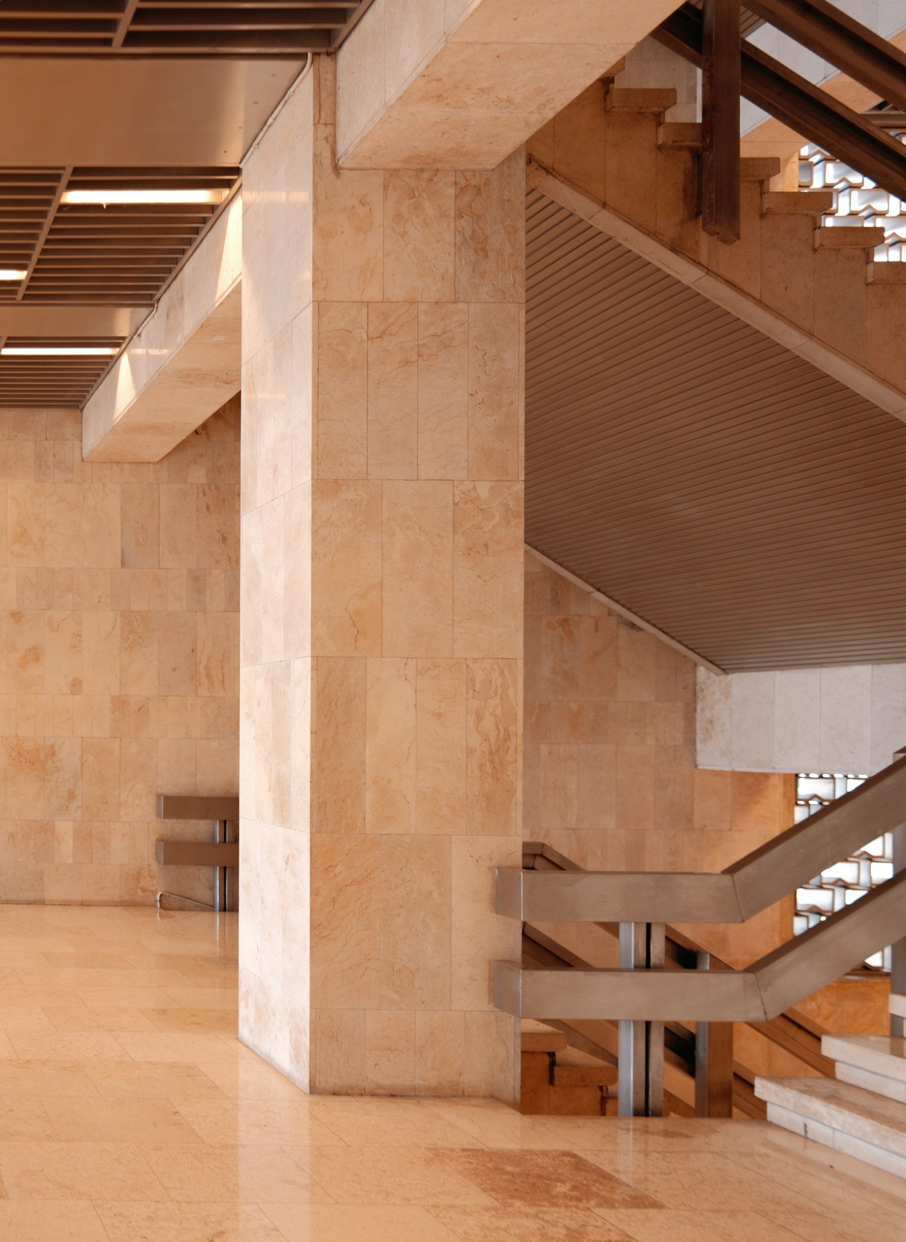 Interior view of a modern building with beige stone walls, columns, and stairs with wooden railings.