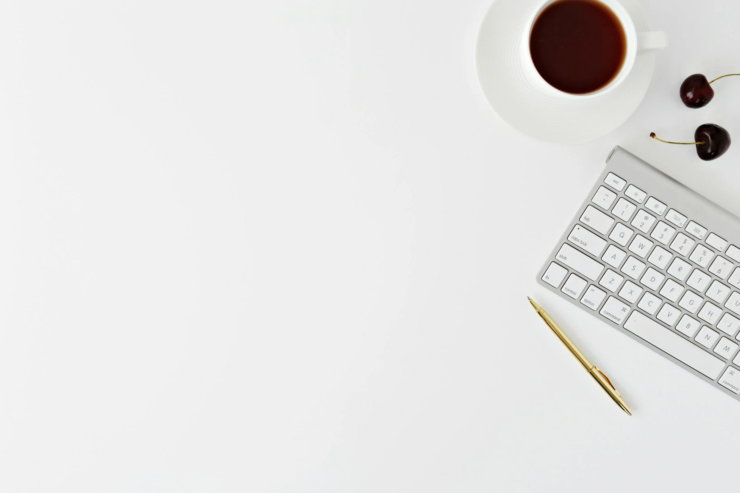 Top view of a white desk with a partial computer keyboard, a gold pen, a white cup of coffee, and two cherries.