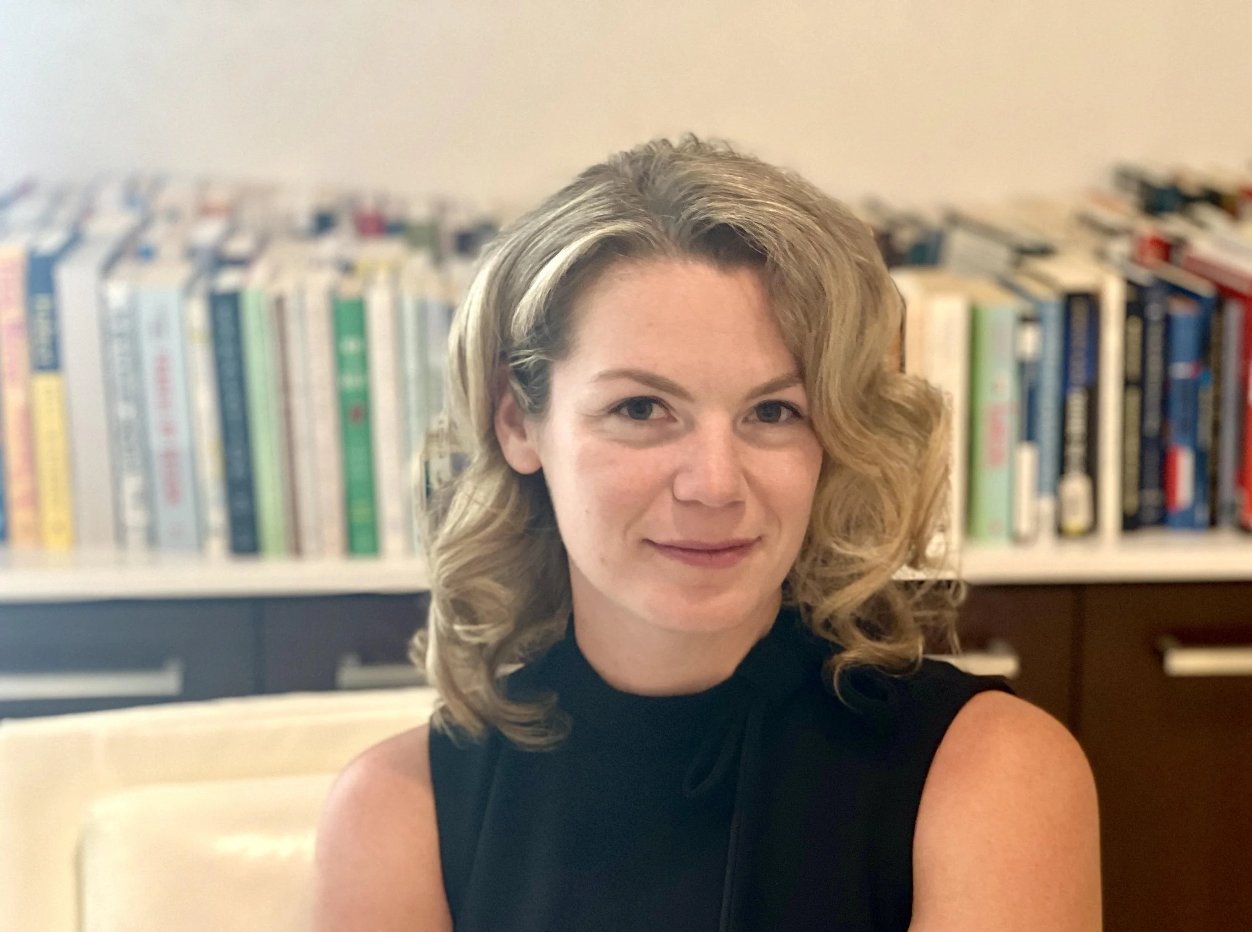 A woman with curly blonde hair wearing a black sleeveless top sitting in front of a bookshelf with numerous books.