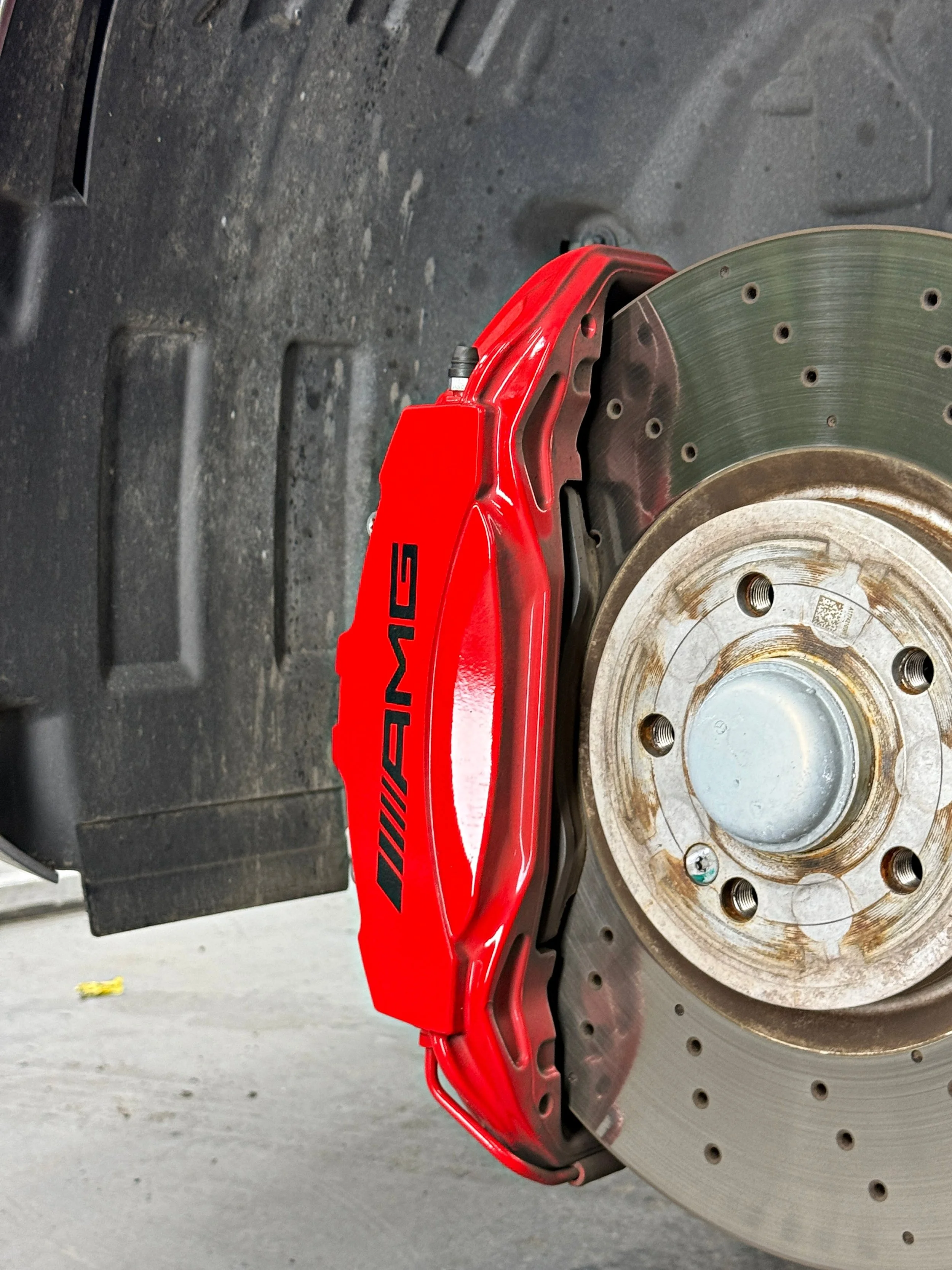 Red AMG brake caliper and wheel arch before cleaning, with dirt visible on the arch liner.