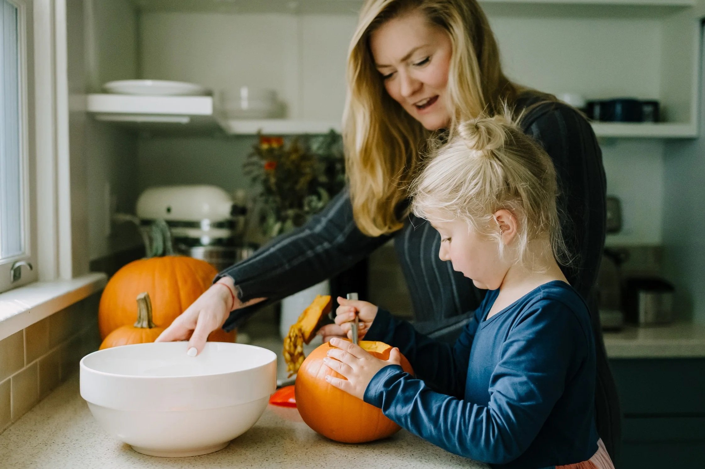 A woman and a girl carving pumpkins in a kitchen, with pumpkins on the counter and white bowls nearby.