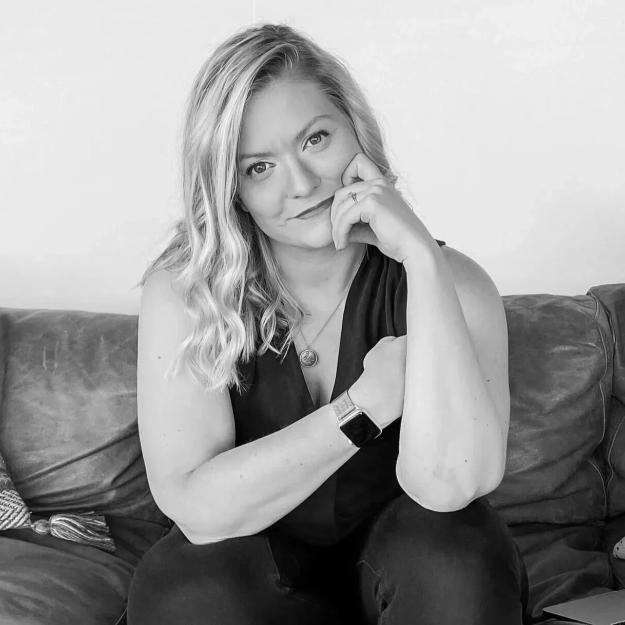 A woman with wavy blonde hair sitting on a couch, resting her chin on her hand, looking at the camera, wearing a black sleeveless top, necklace, and smartwatch.