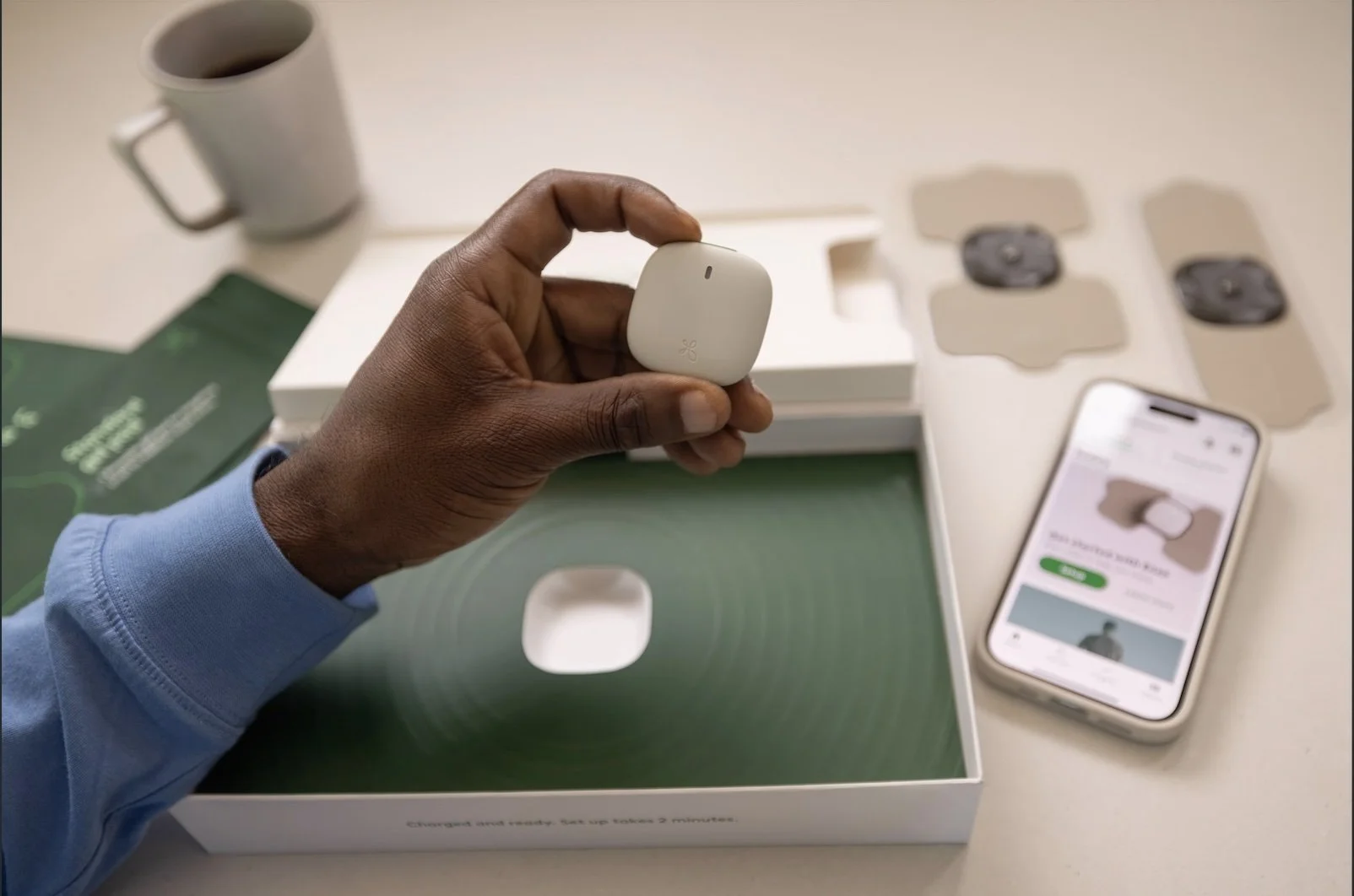 Person holding a white wireless earbud case over an open box with other earbuds and accessories, a smartphone displaying setup instructions, and a coffee mug in the background.