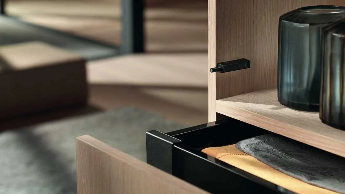 Open wooden cabinet drawer with folded clothes and black container, with shelves above holding black and silver containers.