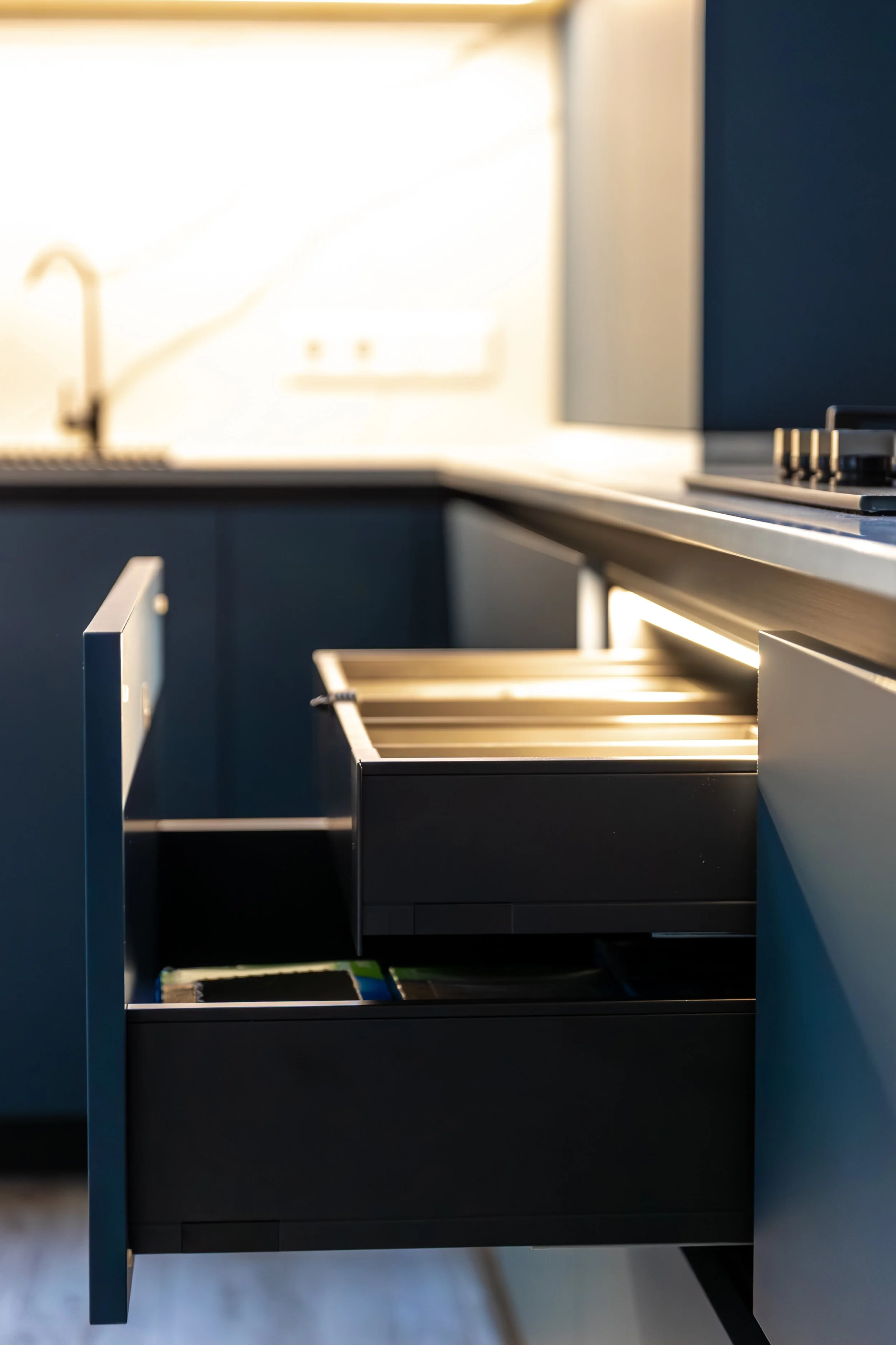 Open kitchen drawer with empty compartments and a view of the kitchen countertop and sink in the background.