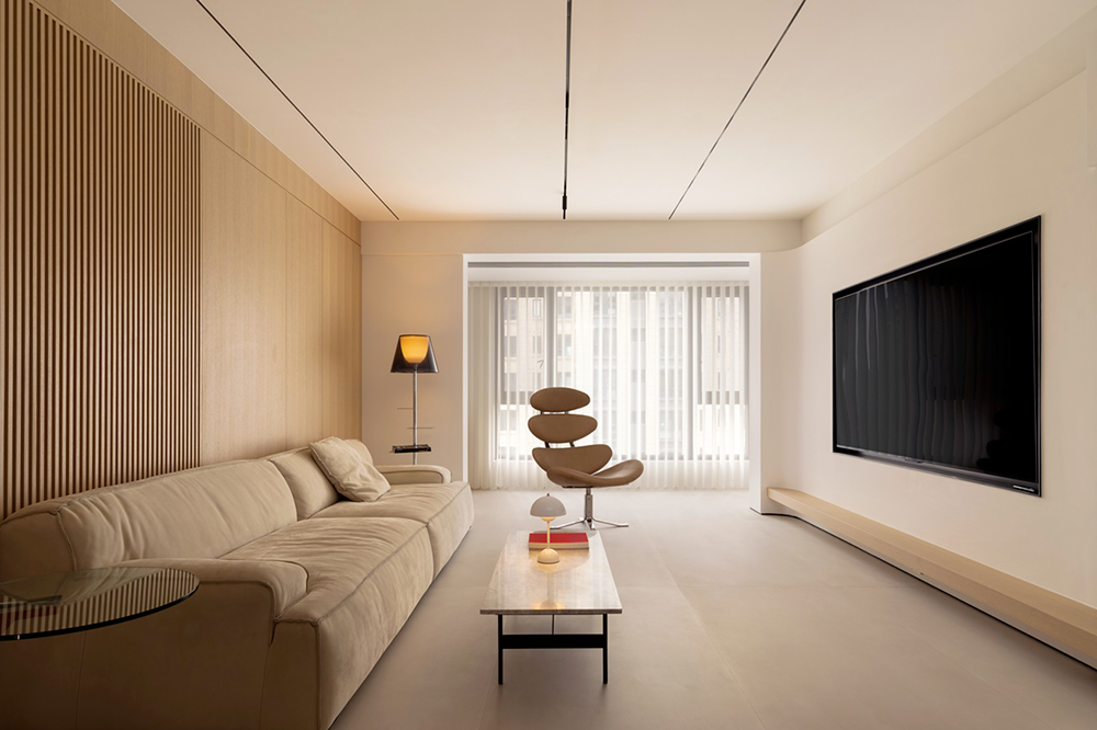Minimalist living room with beige sofa, modern armchair, floor lamp, coffee table with decorative items, large flatscreen TV, and sheer curtains.