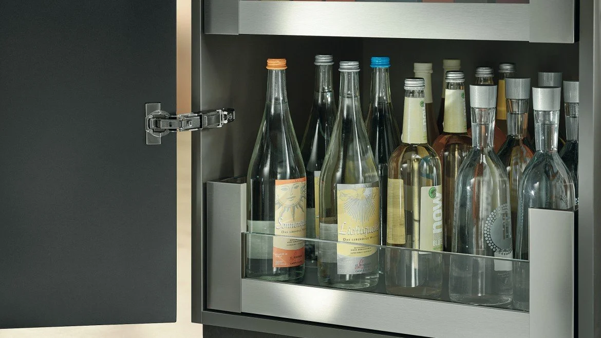 Wine bottles stored inside a dark-colored cabinet with a glass door, some with colored caps and labels.