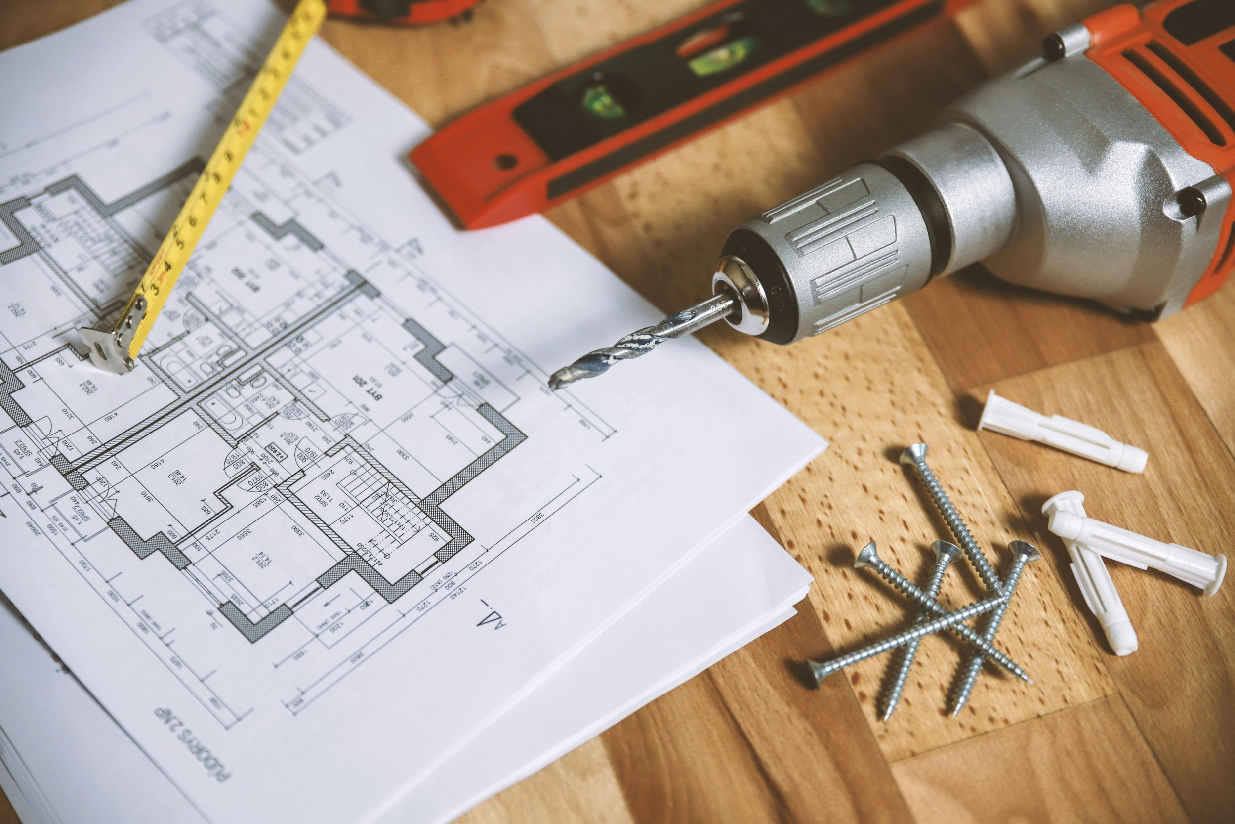Blueprints, a measuring tape, a cordless drill with drill bit, screws, wall plugs, and a level placed on a wooden surface, indicating a home improvement or construction project.