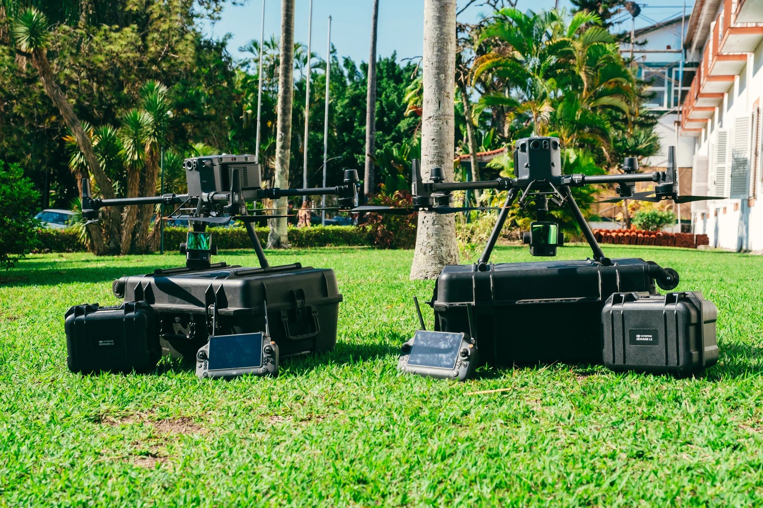 Duas câmeras de drone com controles e cases de transporte posicionados em um gramado em um ambiente ao ar livre, rodeado por árvores e plantas tropicais, com uma casa ao fundo.