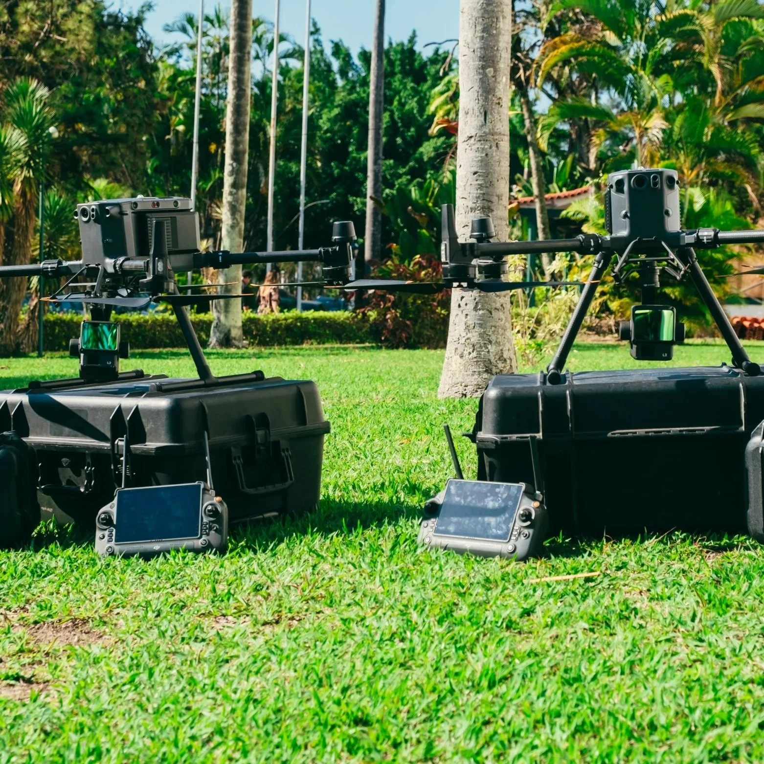 Duas drones grandes apoiados no chão, com controles remotos ao lado, em um parque com grama, árvores e vegetação ao fundo.