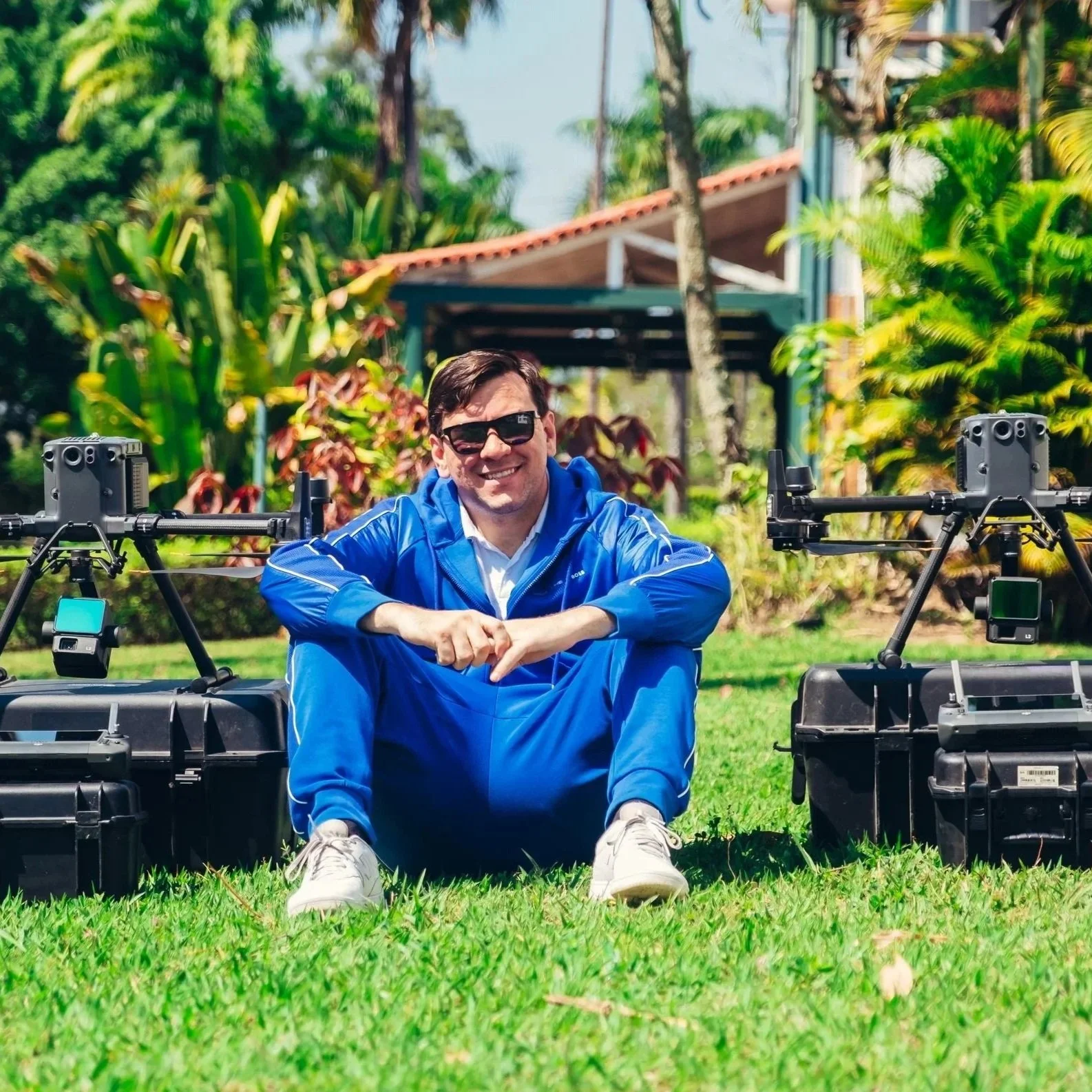 Homem sorridente com óculos escuros, vestindo jaqueta azul, sentado em grama com equipamentos de drones ao seu lado em um jardim com plantas verdes e árvores ao fundo.