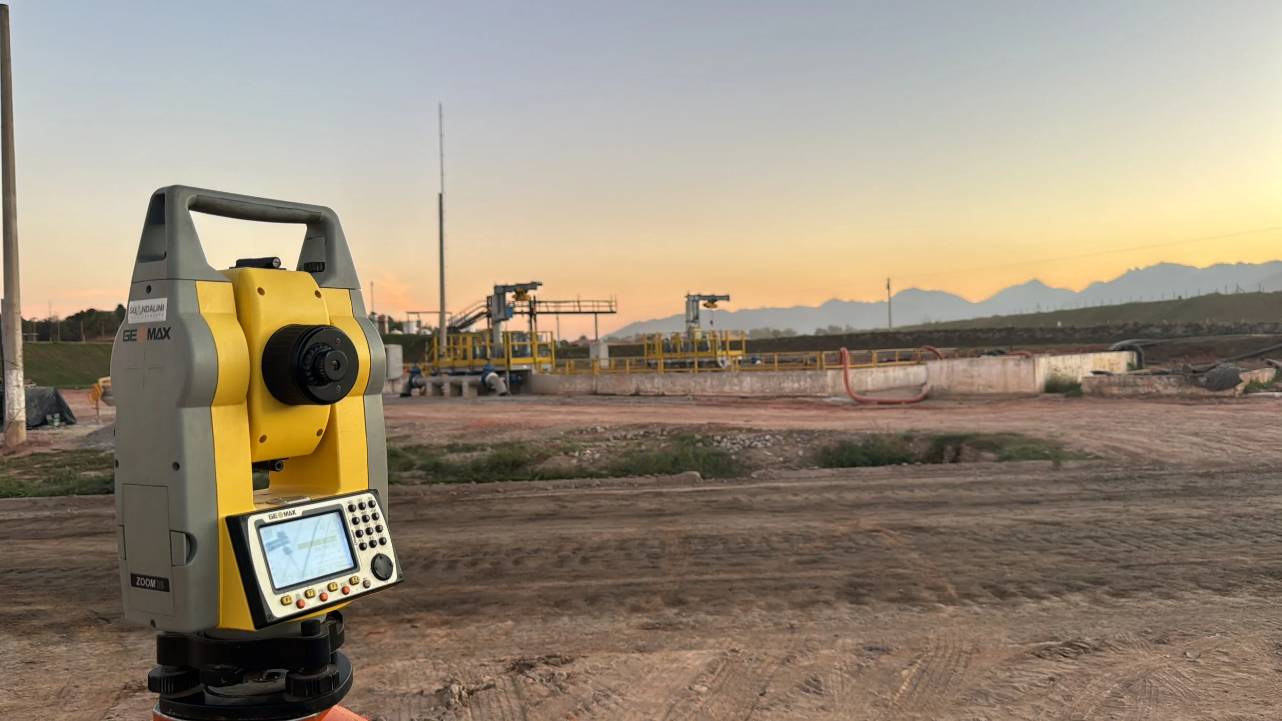 Estação total de topografia em primeiro plano, campo de terra e infraestrutura de petróleo e gás ao fundo, durante o pôr do sol com montanhas ao horizonte