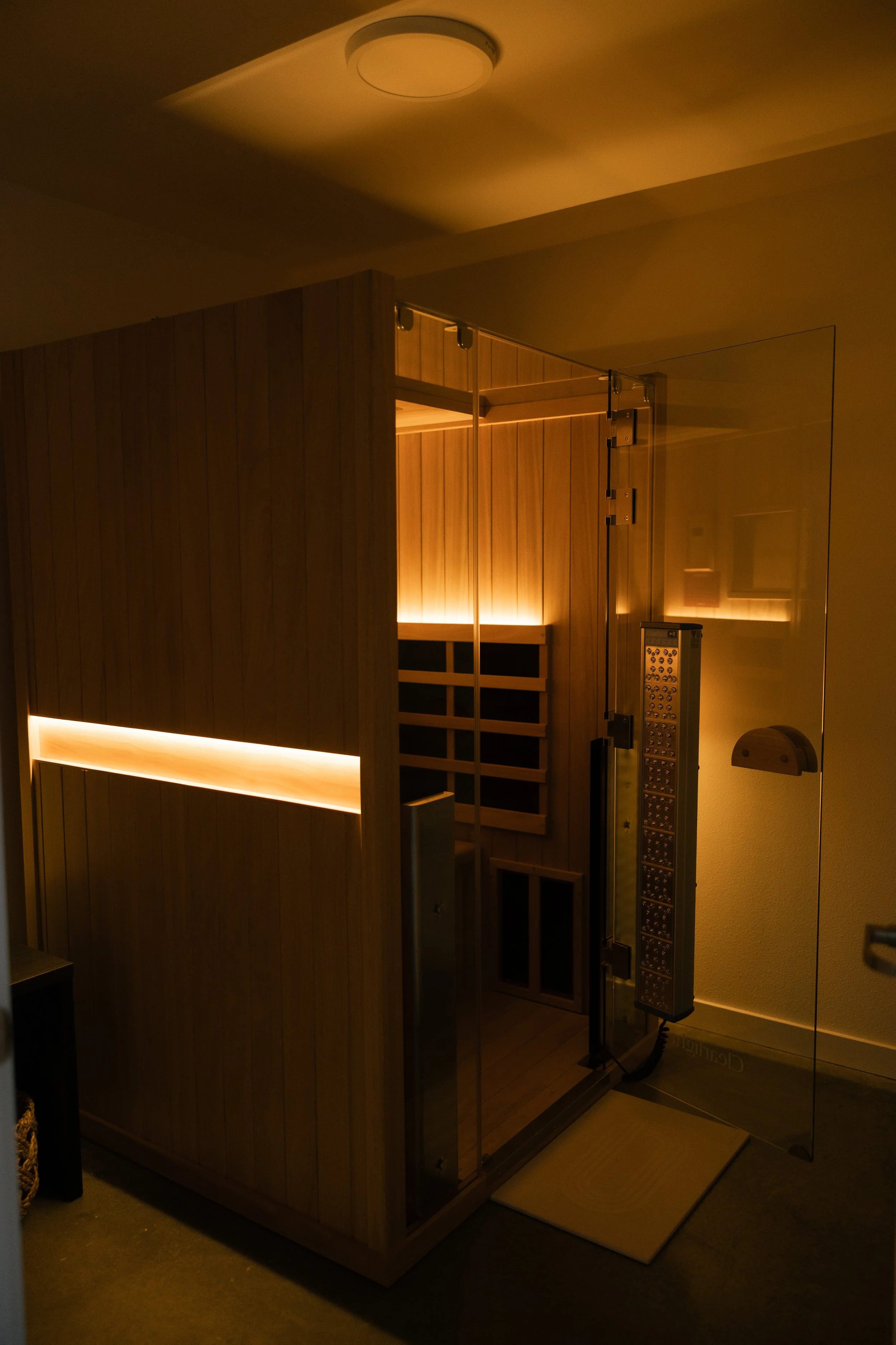 A wooden sauna with a glass door and interior lighting, located in a room with beige walls and a small mat outside the door.