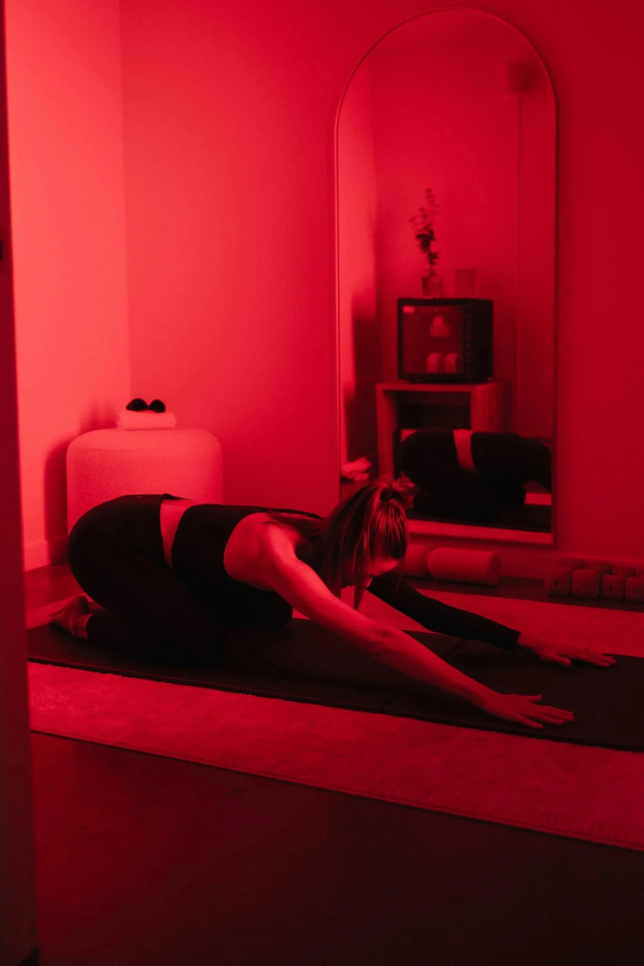 A woman practicing yoga on a black mat in a dimly lit room with red lighting, in a child's pose position, with a mirror reflecting her and the room's decor including a small television, a shelf with vases, and a potted plant.