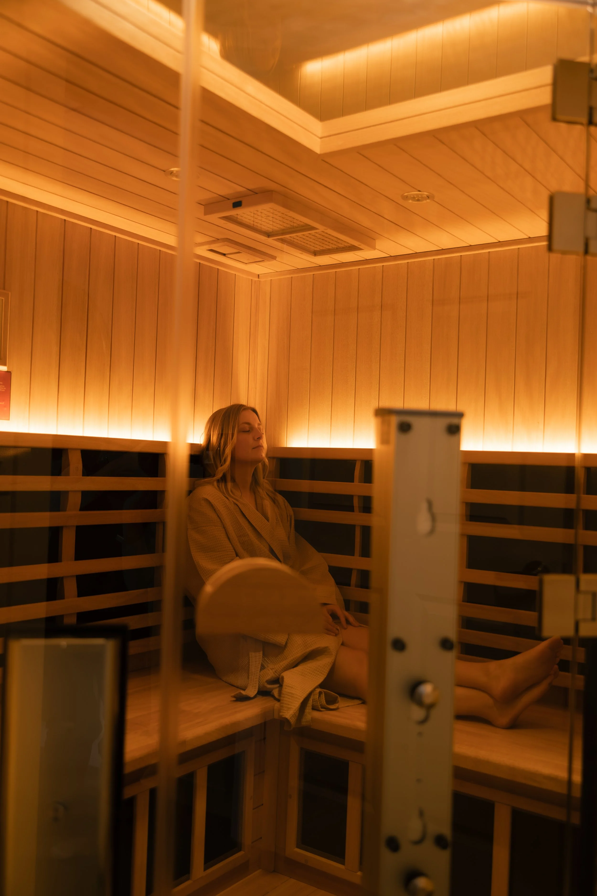 A woman sitting with eyes closed inside a wooden sauna with warm lighting.