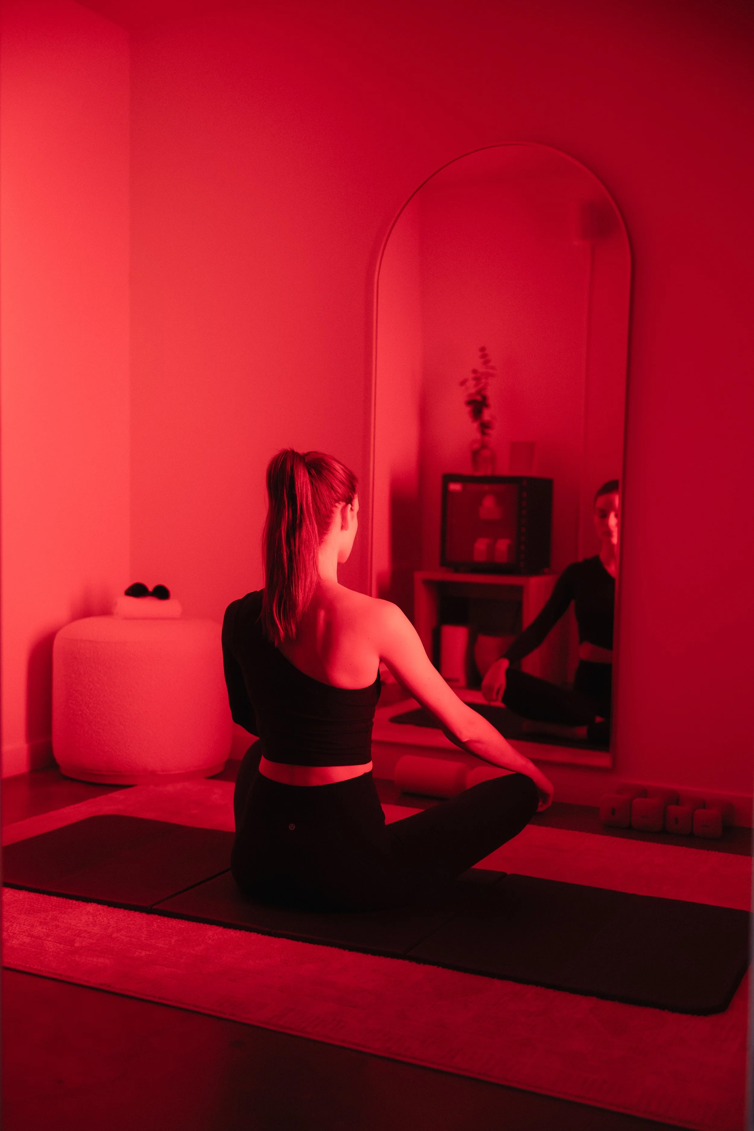 A woman practicing yoga in a dimly lit room immersed in red lighting, sitting on a yoga mat with a mirror behind her, looking at her reflection, with a TV and a small dresser in the background.