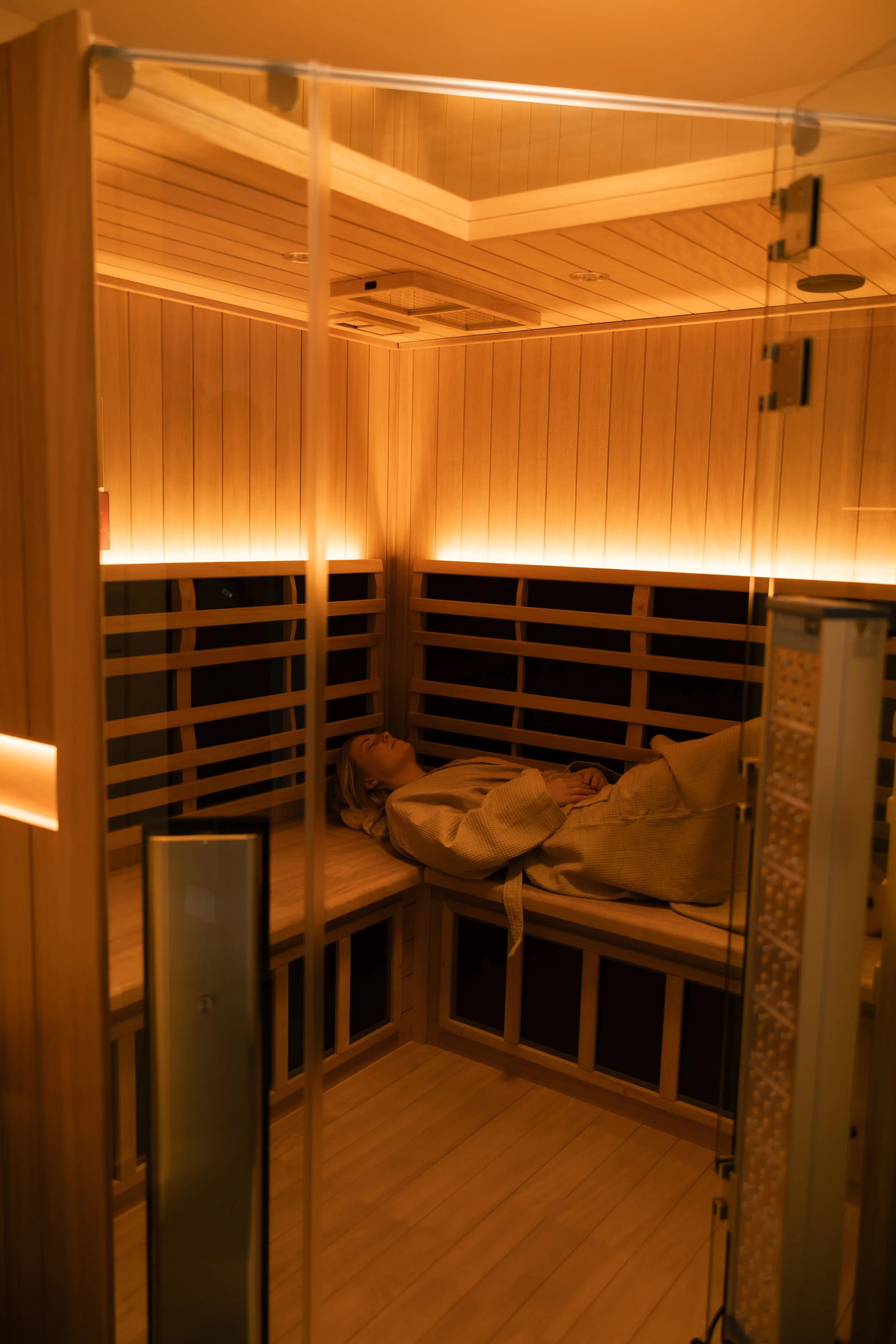 Woman sleeping in a wooden sauna room, lying on a low bed with wooden slat walls and ambient lighting.