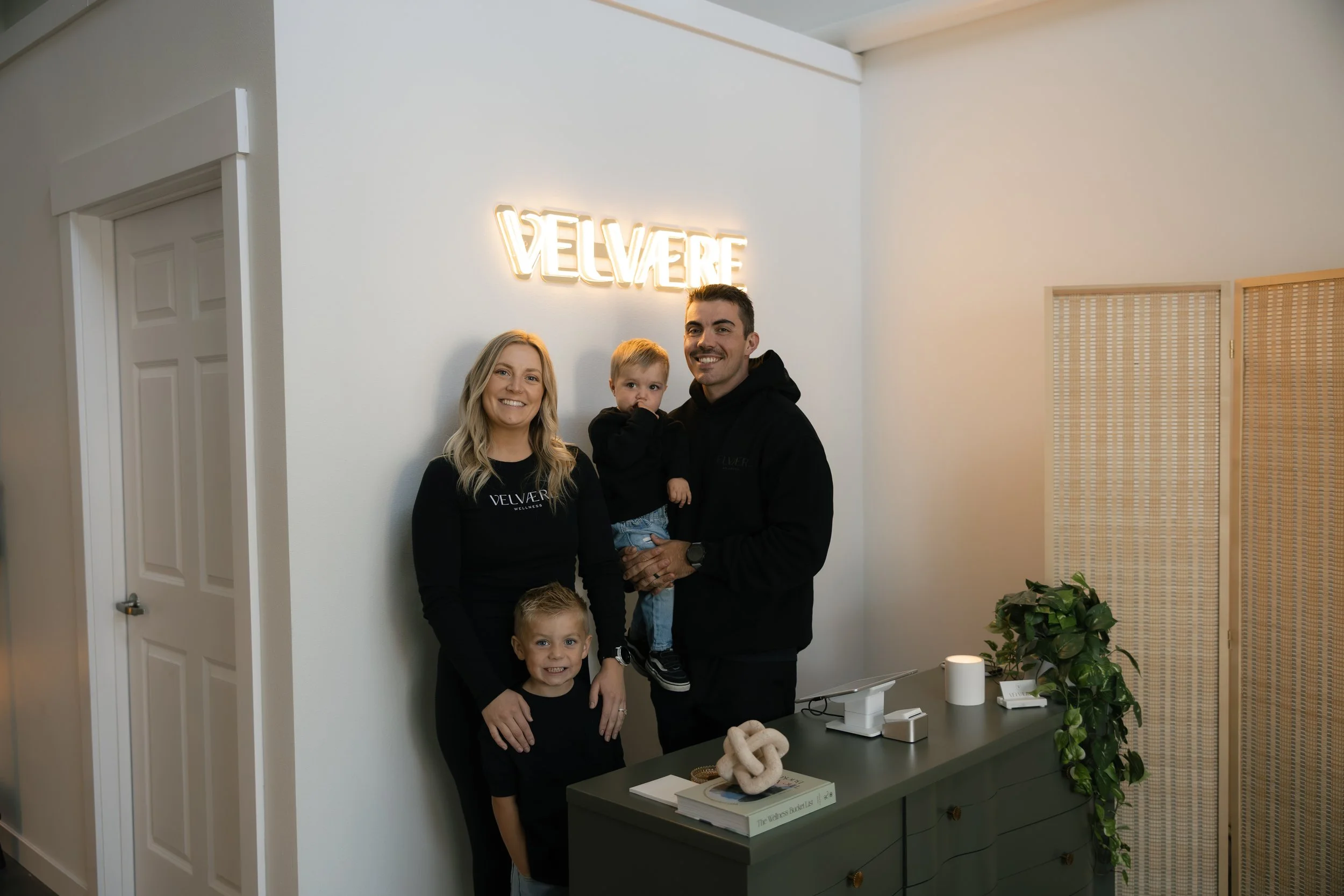 A family of four standing indoors near a wall with a backlit sign. The woman and man are smiling; the woman is wearing a black long-sleeve shirt with 'VELVERE' written on it, and the man is holding a small child. The small boy in front is smiling, and there is another child partially visible at the bottom of the frame. There is a green dresser with books, a decorative sculpture, and a potted plant on it, and a beige window shade is seen in the background.