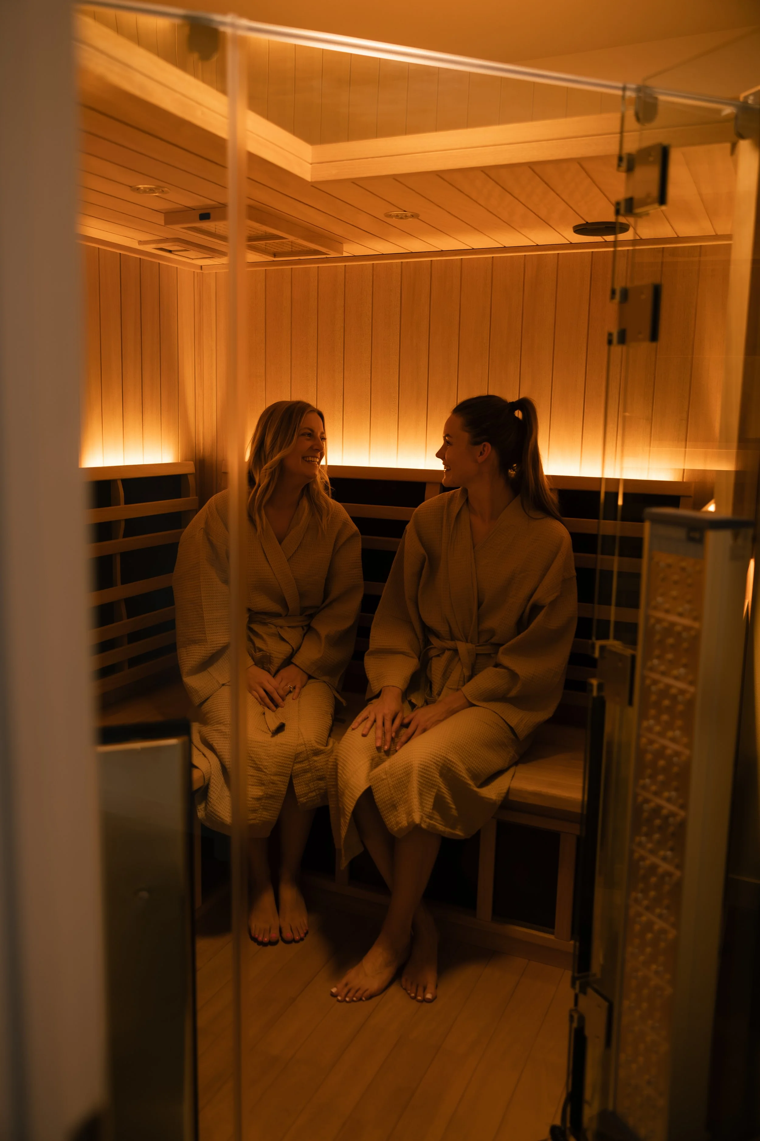 Two women sitting inside a wooden sauna, smiling at each other, wearing beige robes.