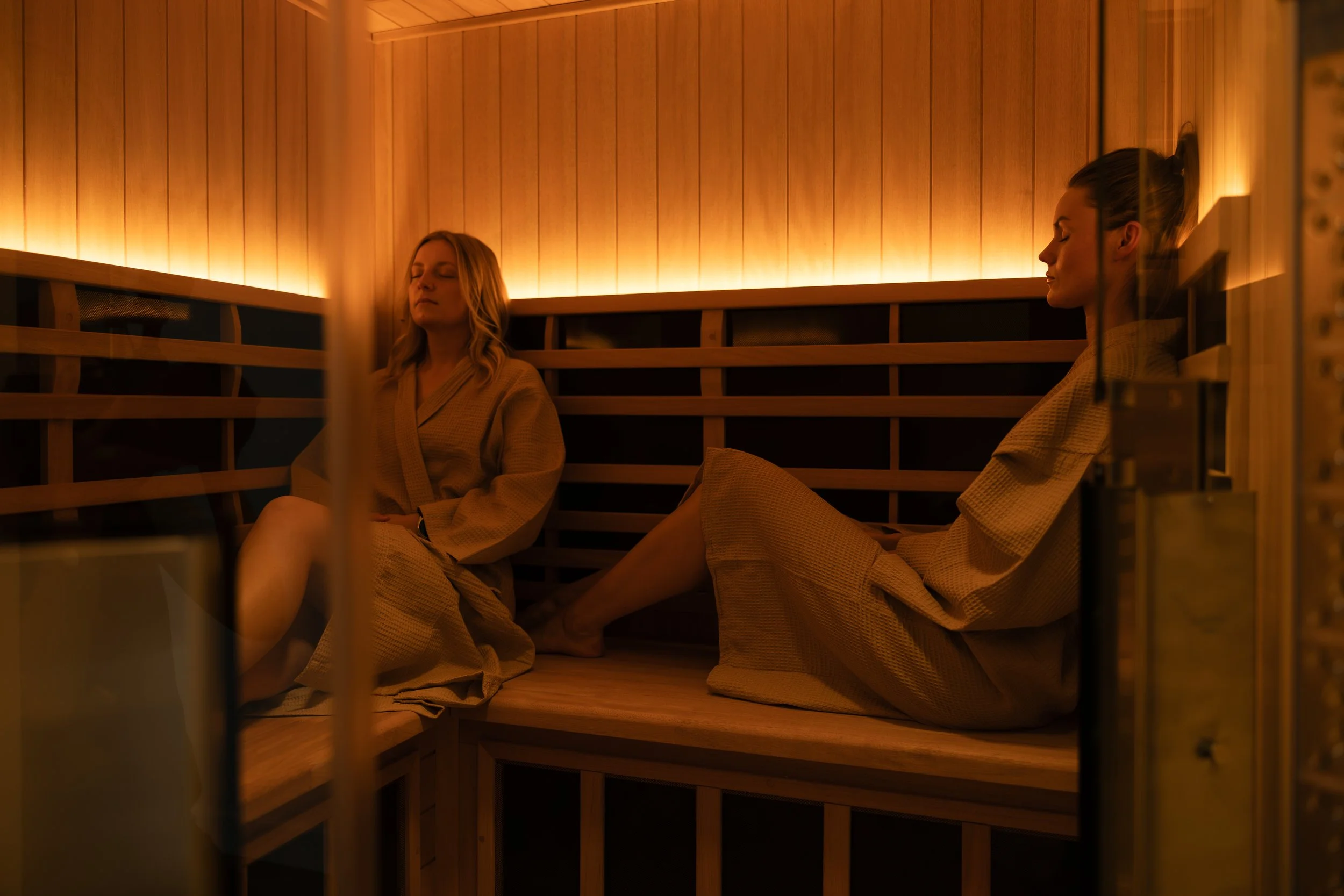 Two women sitting in a sauna with closed eyes, wearing beige robes, surrounded by wooden walls and benches, illuminated by warm lighting.