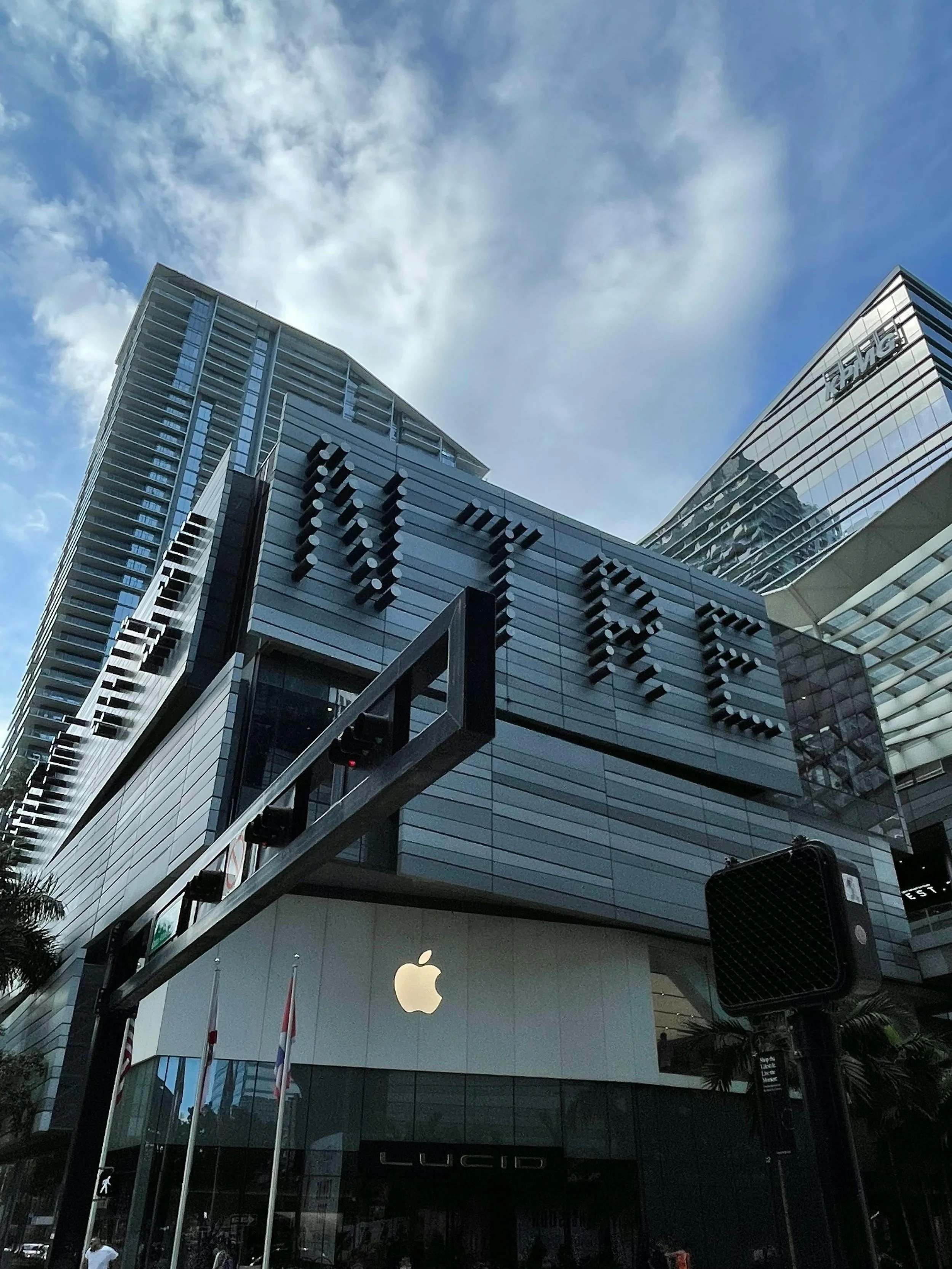 Exterior of Apple Store in a modern building with large Apple logo, surrounding high-rise buildings, and a cloudy sky.