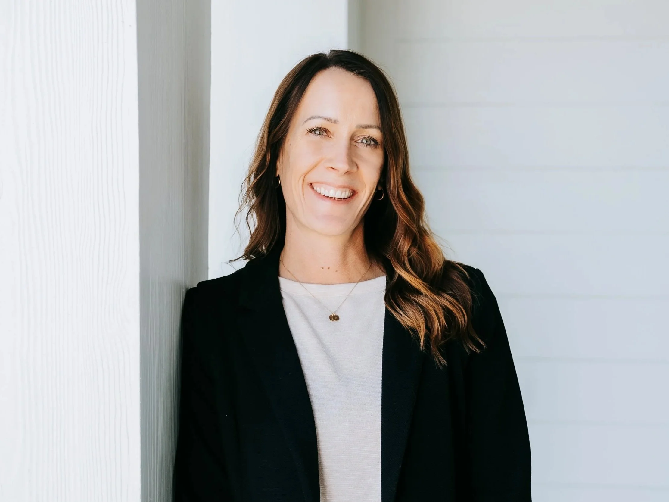 Smiling woman with wavy brown hair, wearing a black blazer and white top, standing against a white wall.