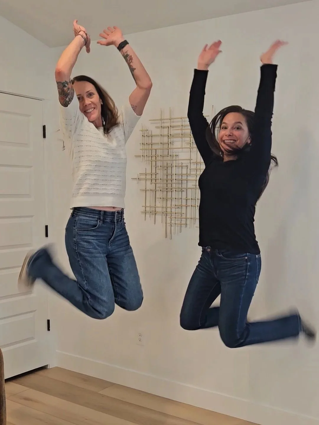 Two women are joyfully jumping in the air indoors, smiling with their arms raised. One woman has long hair, tattoos, and is wearing a white top and jeans, while the other has shoulder-length dark hair and is dressed in a black top and jeans. A geometric wall sculpture is visible in the background.