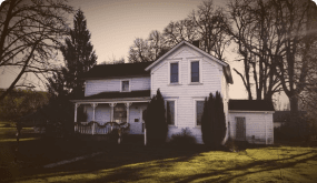 A large white two-story house with a front porch, situated on a grassy lawn with trees in the background during sunset.