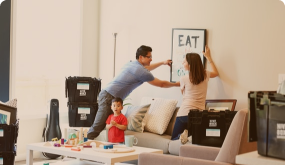 A man and woman are hanging a framed picture on the wall in a living room. A young girl stands nearby watching. The room has various boxes and furniture, suggesting a recent move or renovation.