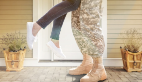 Person in camouflage uniform lifting another person in casual clothing by the legs outside a house with potted plants