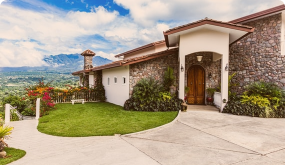 Exterior of a house with stone and stucco walls, a wooden front door, a curved driveway, and landscaped lawn with bushes and flowers.