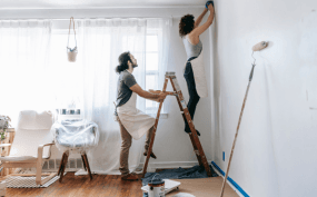 Two women decorating a wall in a bright living room. One woman is on a ladder, sticking wallpaper or artwork, while the other supports her. The room has large windows, a chair, and a floor lamp.