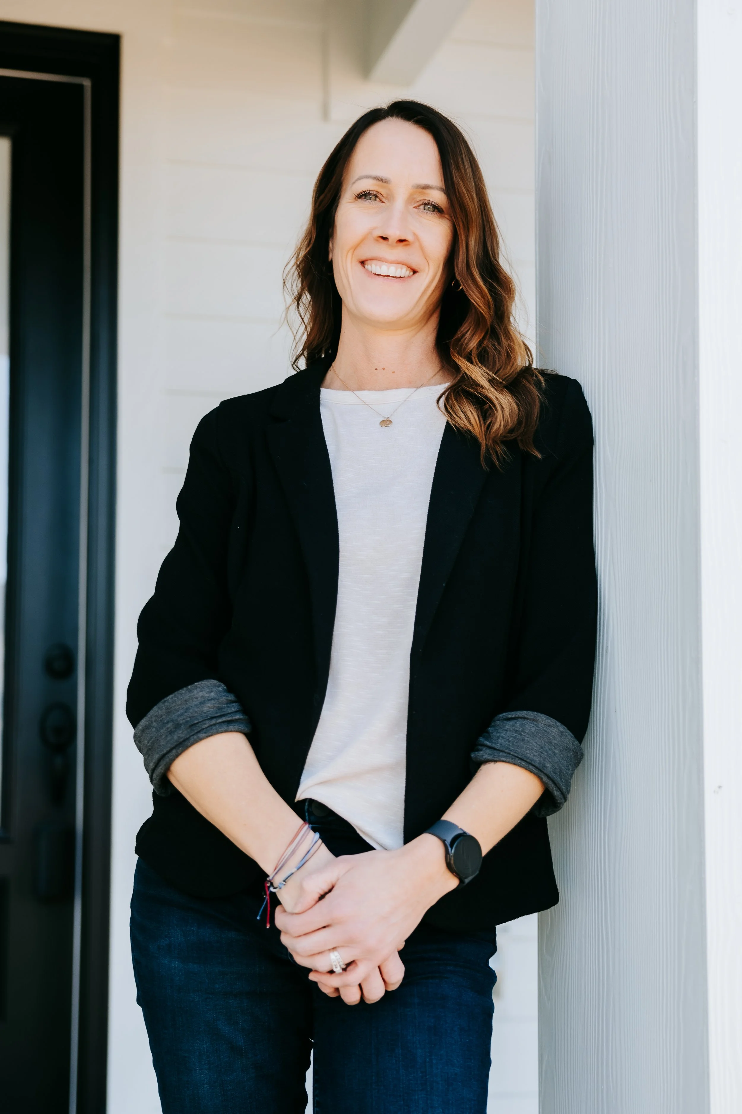 A woman with shoulder-length brown hair wearing a black blazer, white t-shirt, and dark jeans, standing outdoors and smiling.