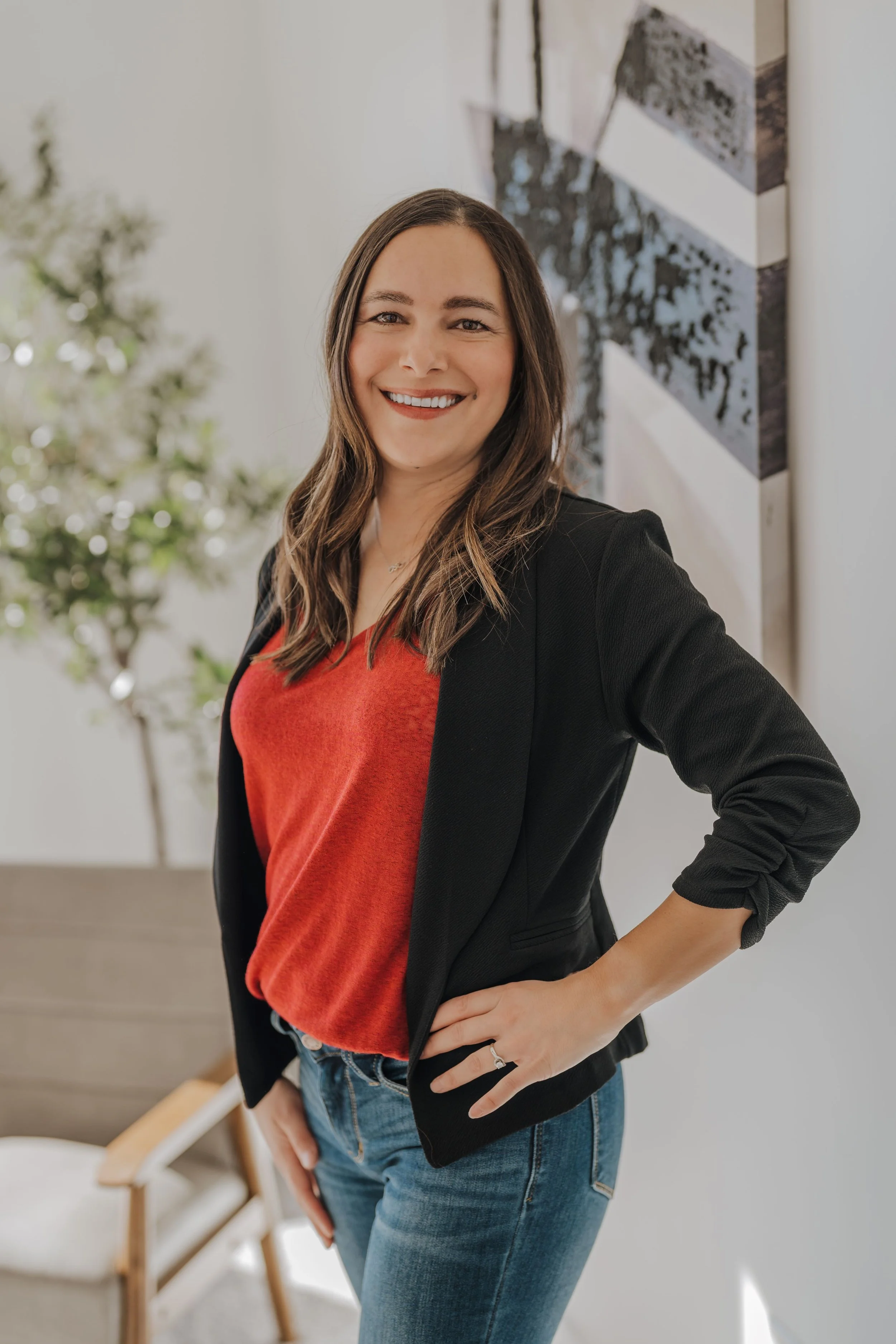 A smiling woman with long brown hair wearing a red shirt, black blazer, and blue jeans, standing with her hand on her hip indoors with a blurred background of a plant and abstract art on the wall.