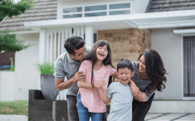 A family of four has fun outside in front of a house, with two children laughing and parents smiling and playing with them.