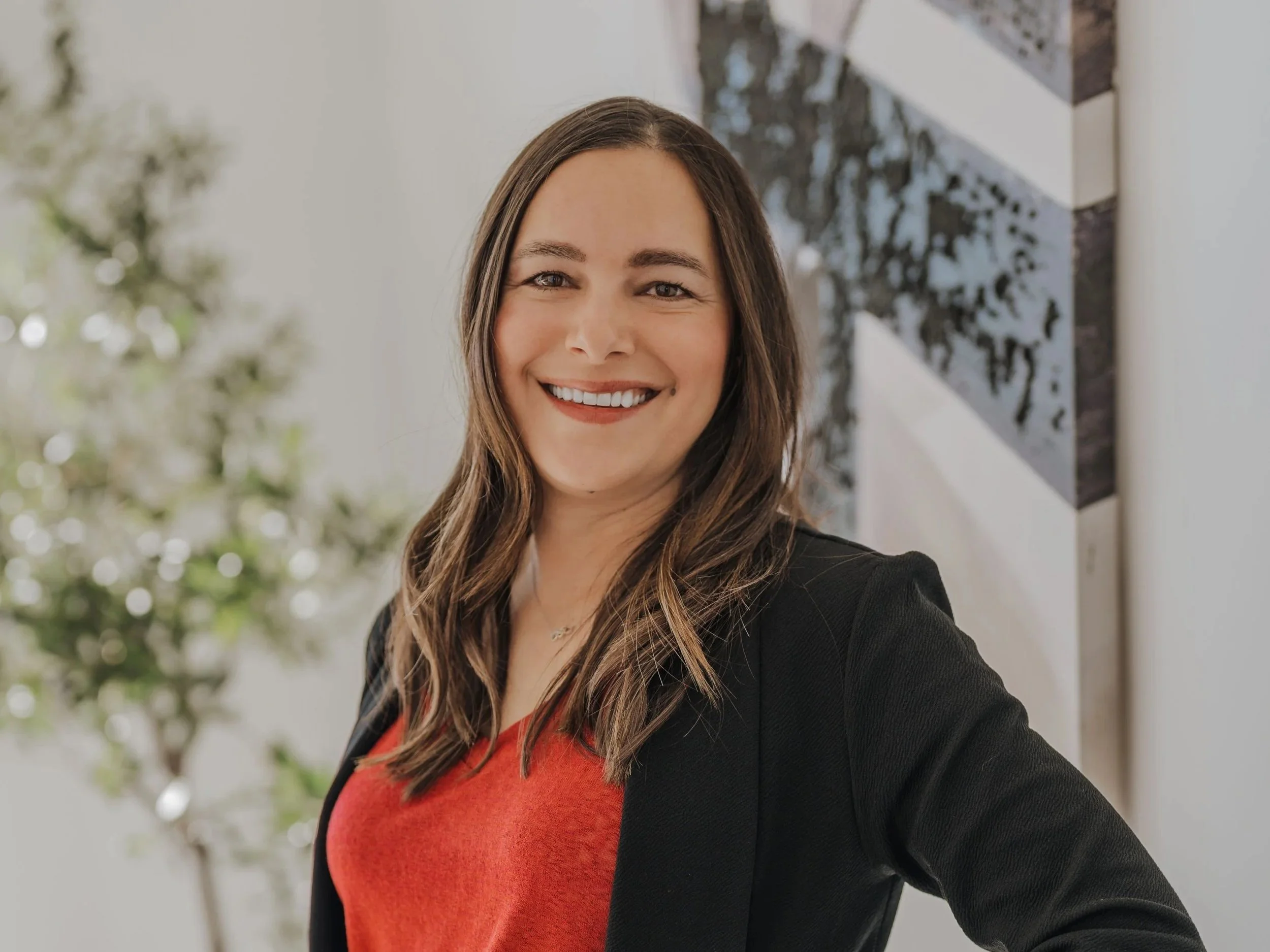 Portrait of a woman with long brown hair smiling and wearing a black blazer over an orange top, standing indoors near a blurred background with a plant and artwork.