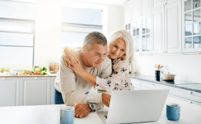An elderly couple sitting together in a bright kitchen, looking at a laptop screen and smiling.