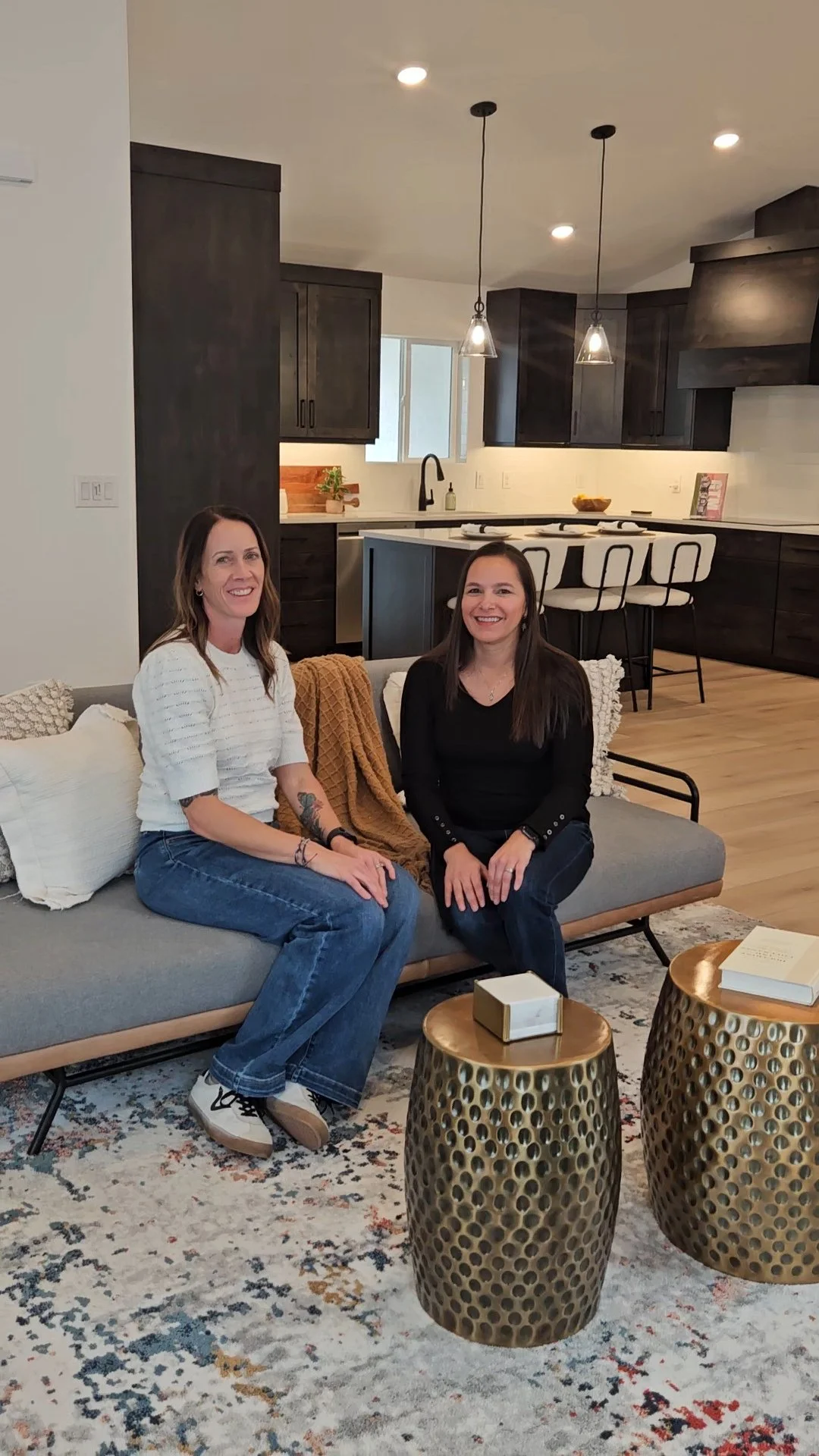Two women sitting on a modern gray sofa in a living room with a kitchen in the background, featuring dark cabinets, a window, and pendant lights, with two metallic gold side tables in front.