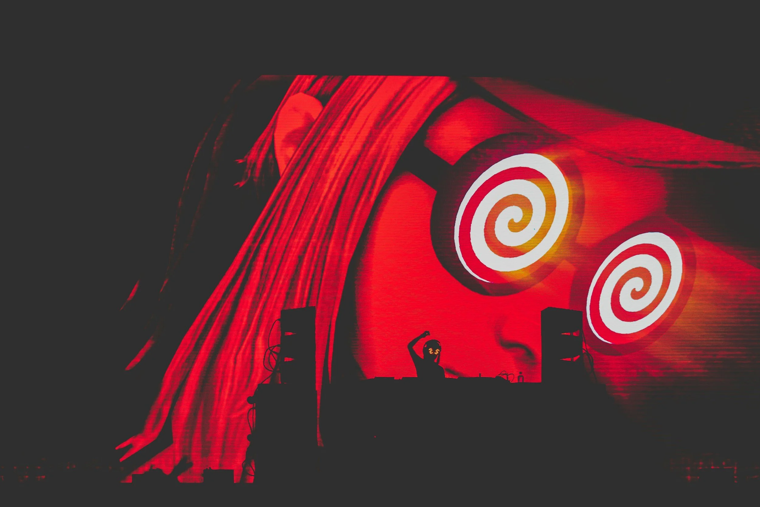 Pantalla de cartelera con imágenes en tonos rojos y blancos, mostrando círculos en espiral y una figura de un DJ en la parte inferior.