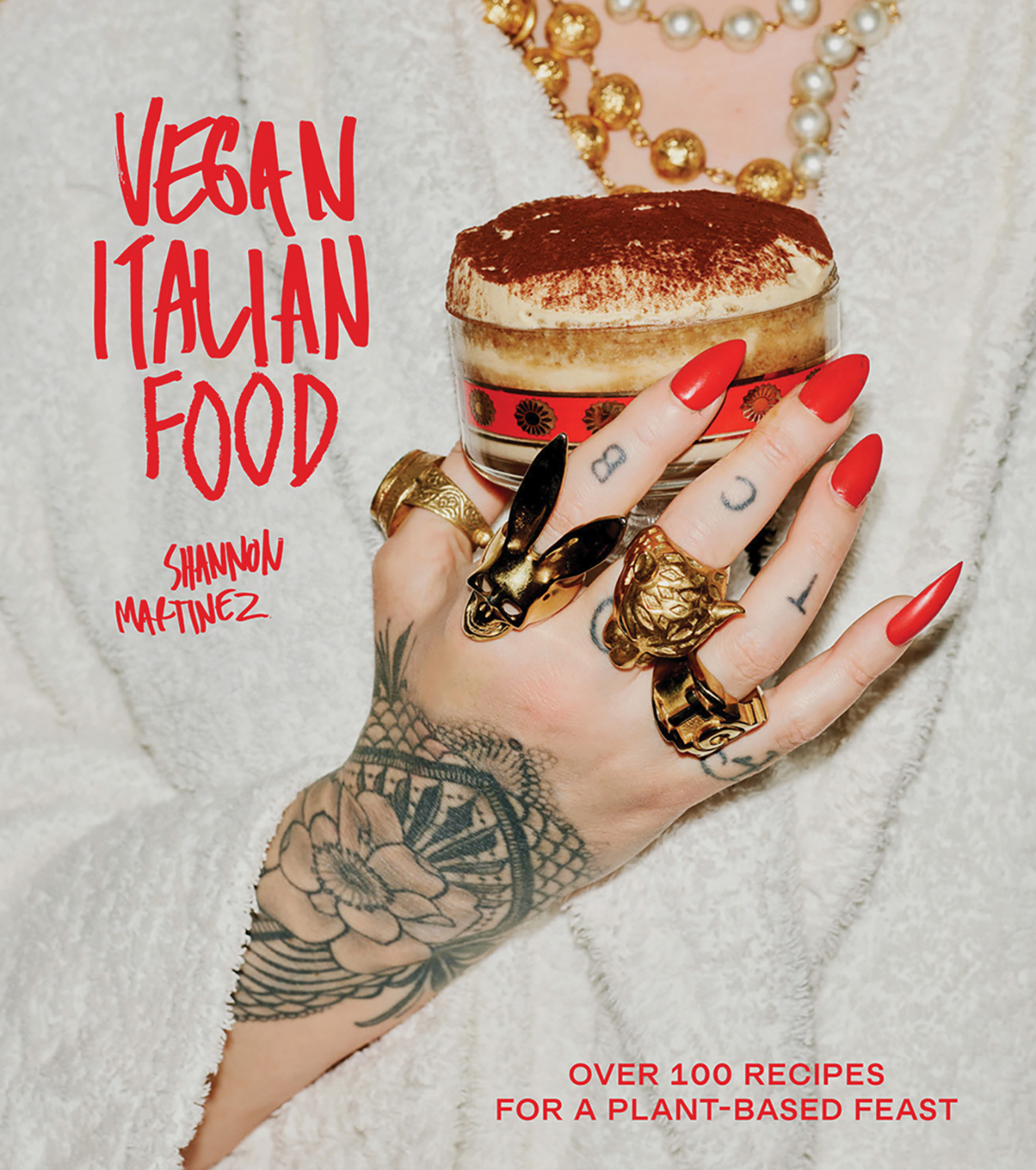 Book cover for Vegan Italian Food. A person holding a glass of tiramisu topped with cocoa powder, with long red nails, multiple rings, and tattoos on their hand and arm. They are wearing a pearl and gold bead necklace and a fuzzy white dressing gown.