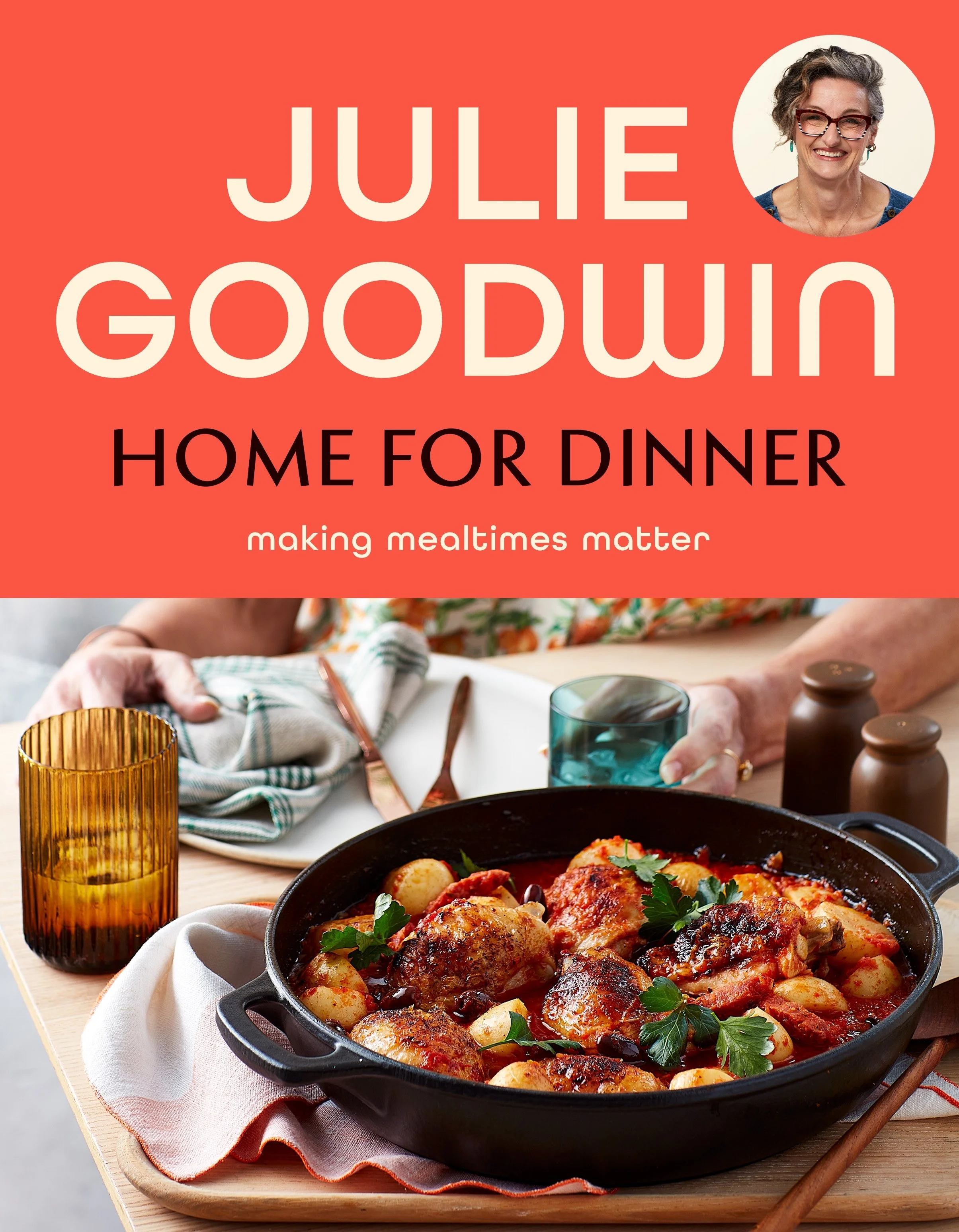 A close-up of a cast iron skillet filled with roasted chicken and gnocchi in a tomato sauce, garnished with fresh parsley, on a wooden table with a white cloth, surrounded by amber glassware and brown bottles, and a woman in the background preparing a meal.