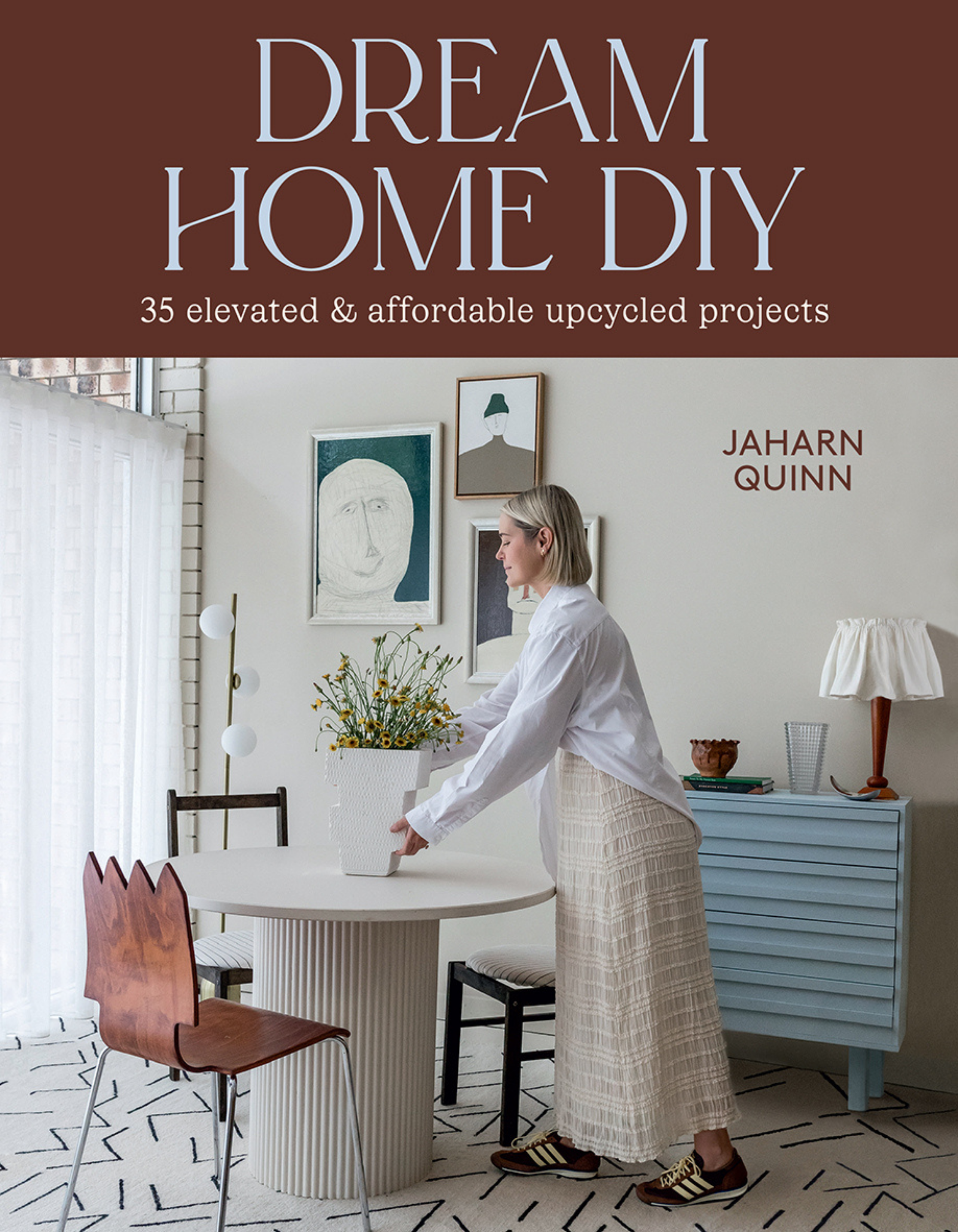 Book cover for Dream Home DIY, featuring a photo of a woman wearing a white blouse, beige plaid skirt, and sneakers placing a white flowerpot on a dining table in a bright, modern kitchen or dining room.