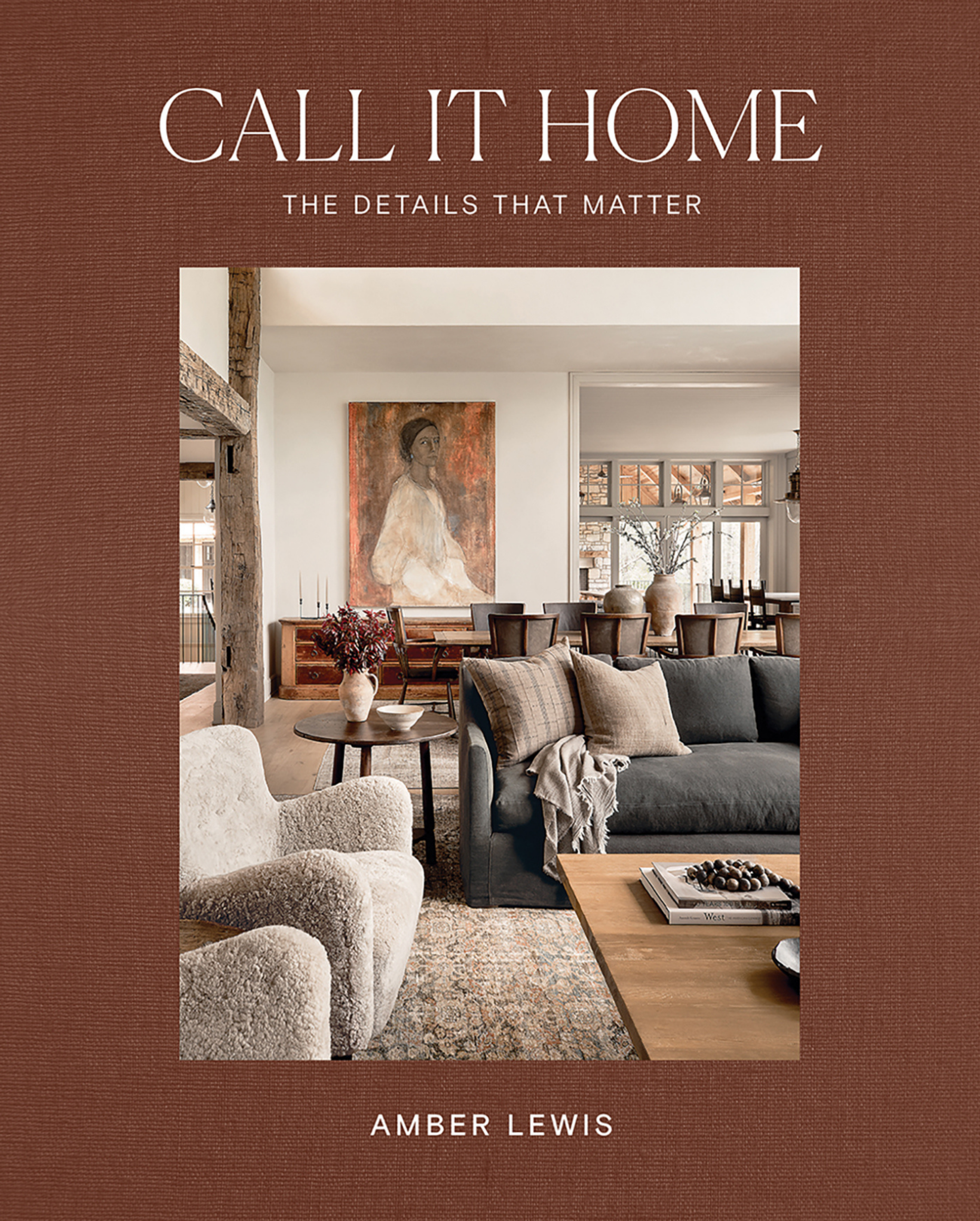 Book cover titled "Call It Home: The Details That Matter" by Amber Lewis, featuring a photo of a cozy, modern living room with a blue sofa, a wooden coffee table with a book and decorative balls, a patterned rug, a large painting of a woman, and a dining area in the background.