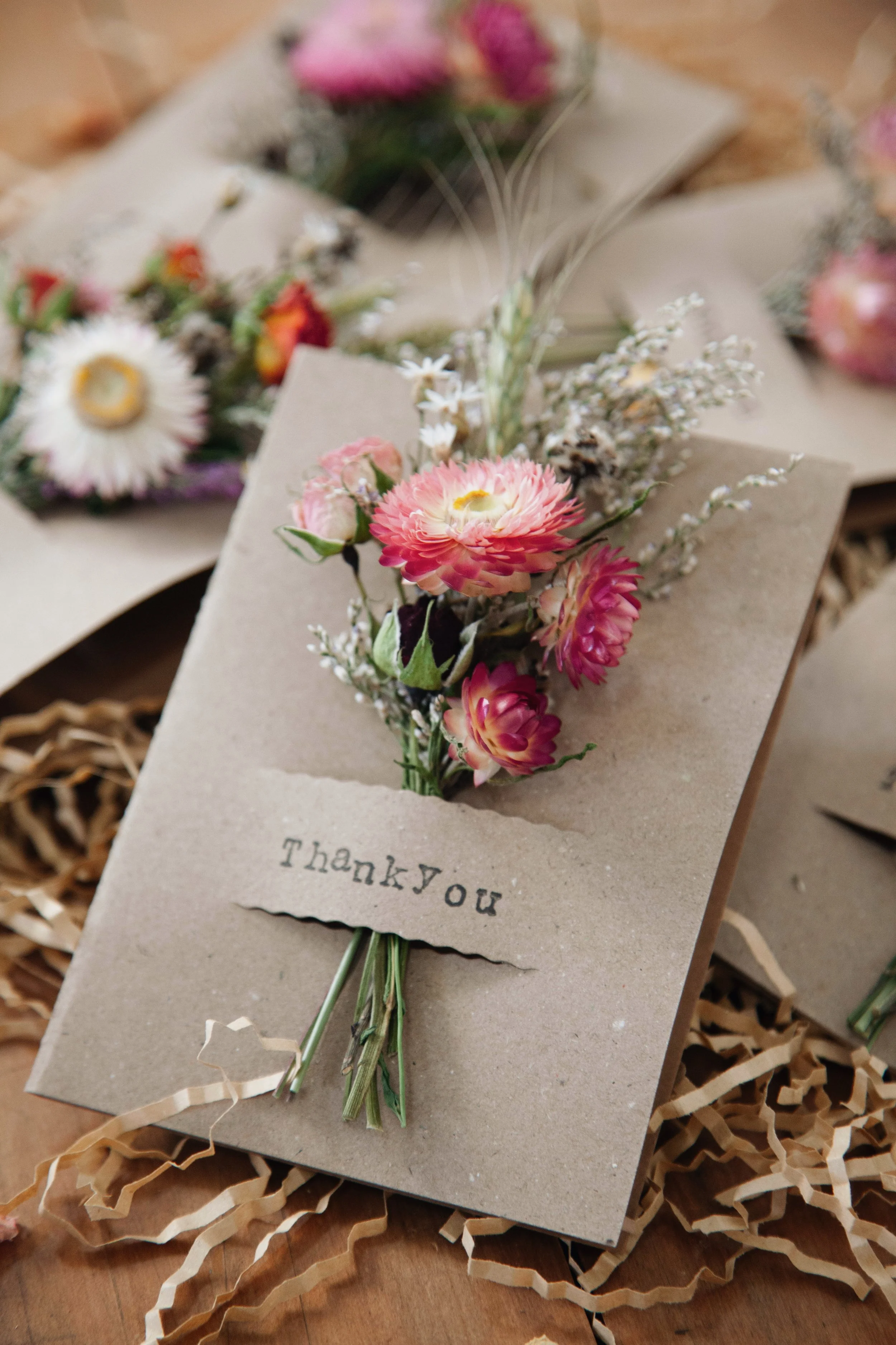 A small bouquet of pink and white flowers with greenery, wrapped with a 'Thank you' note, resting on shredded paper or kraft paper.