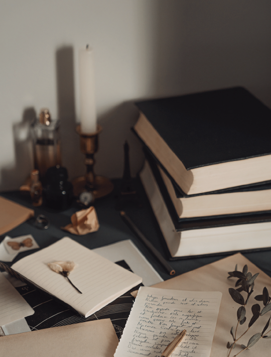 A desk with three stacked black books, a notepad with handwritten notes and a pen, a candle, papers, and small decorative objects, with a branch of eucalyptus leaves nearby.