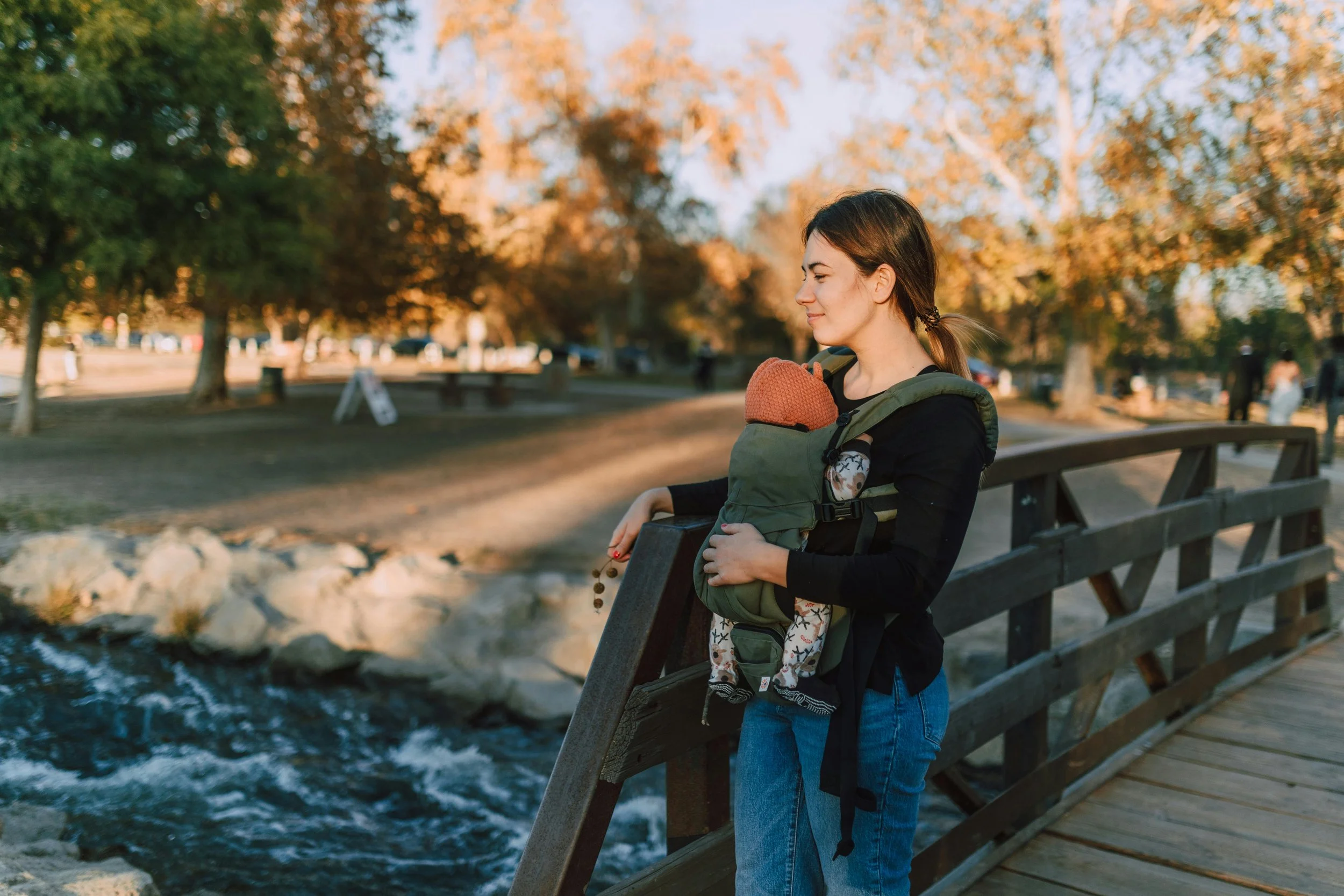A woman with brown hair in a ponytail, wearing a black top and blue jeans, standing on a park bridge with a creek below, holding a child in a green baby carrier in front of her. The child is dressed in patterned clothing. The scene is set in autumn, with trees in the background displaying fall colors and a few people in the distance.