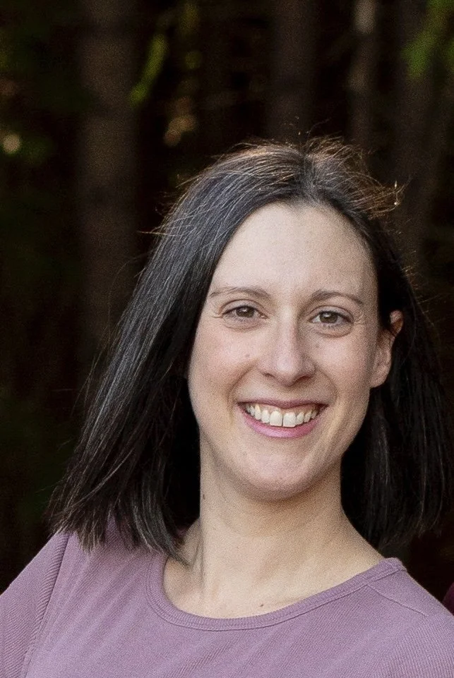 Smiling woman with shoulder-length dark hair wearing a mauve top, outdoors with blurred trees in the background.
