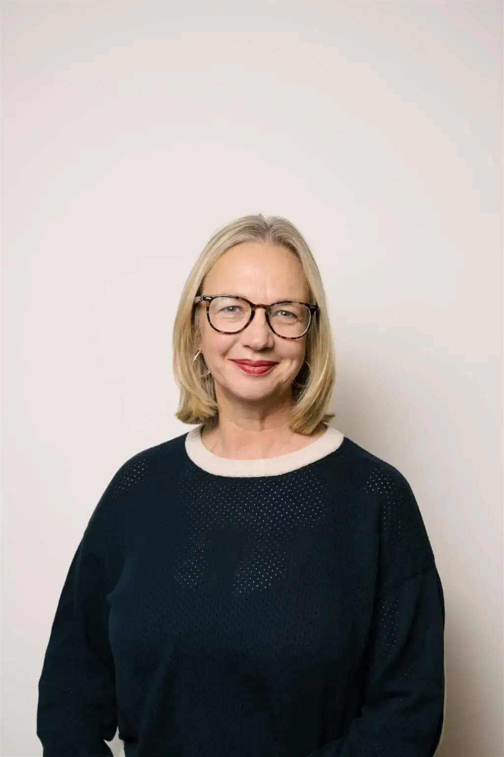 A middle-aged woman with blonde hair and glasses, wearing a black sweater with a white collar, standing against a plain off-white wall.