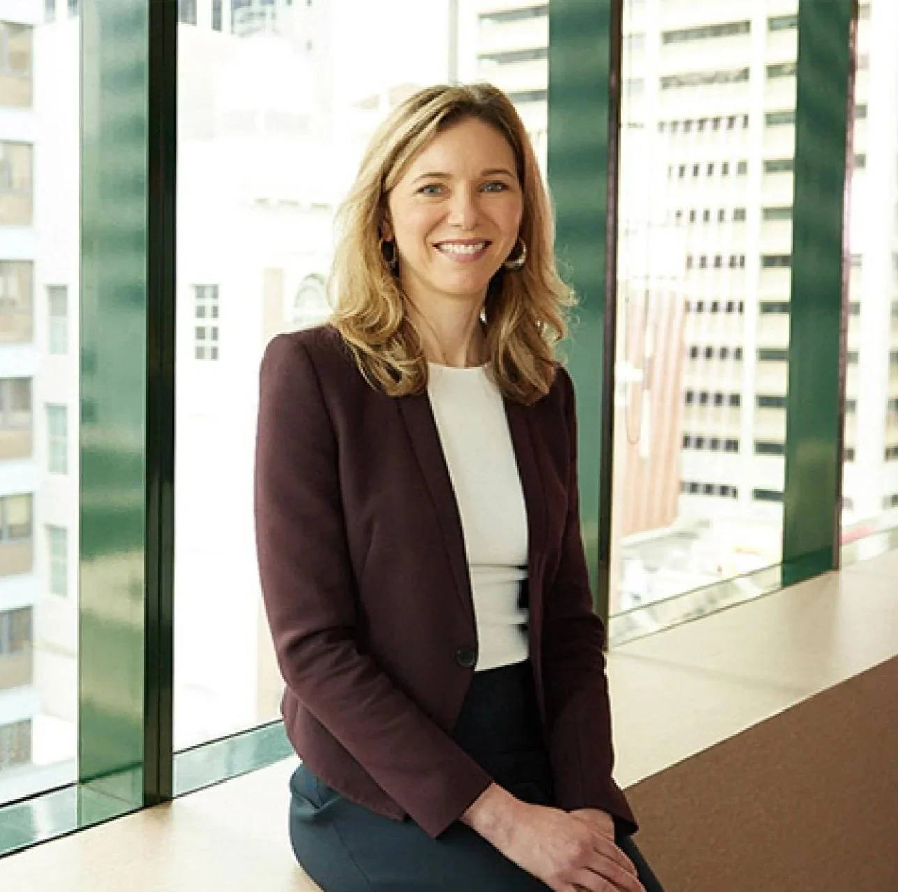 A woman with shoulder-length blonde hair, wearing a burgundy blazer and white top, smiling while sitting by a large window in a high-rise building with cityscape in the background.