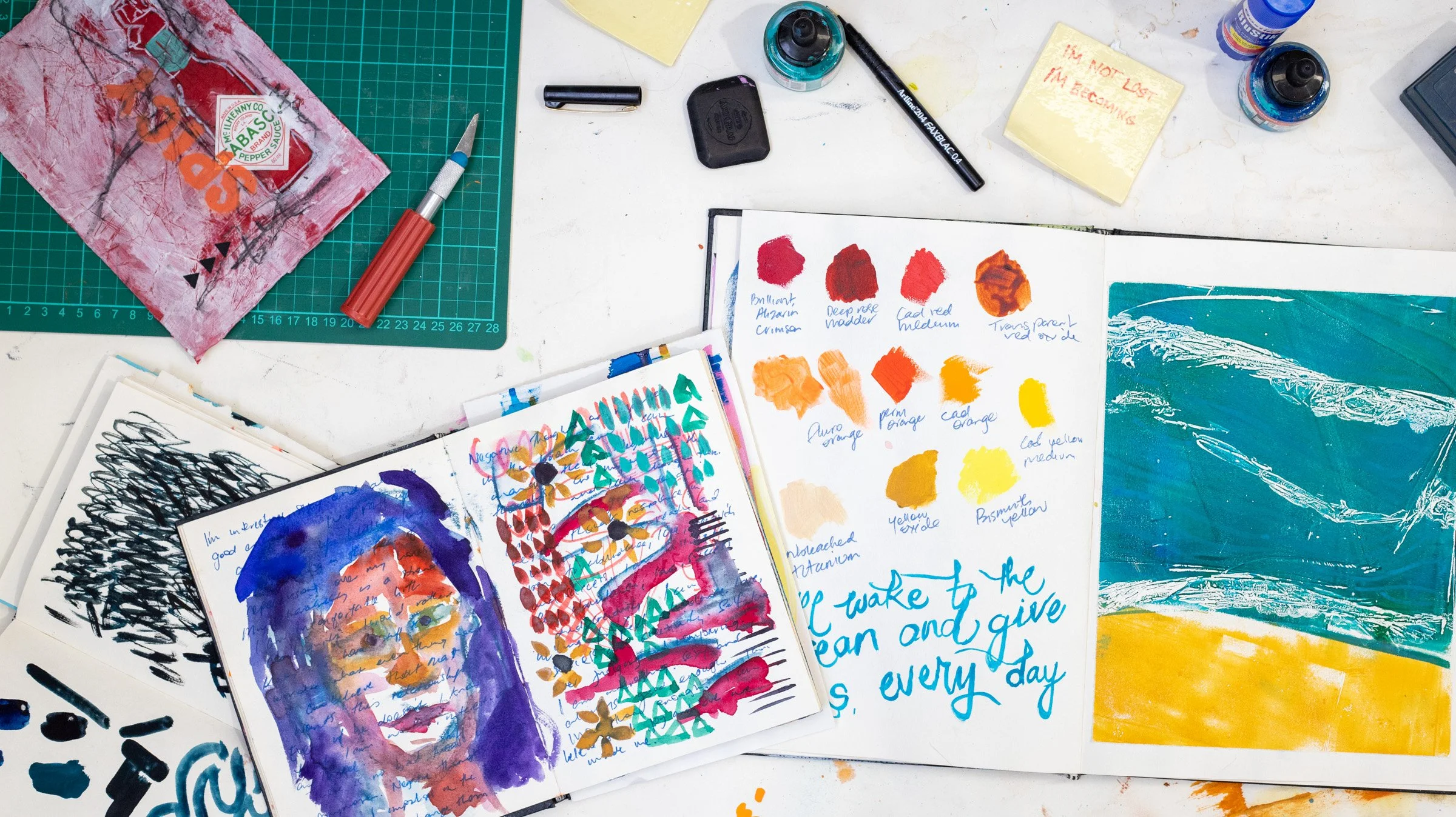 An art workspace with watercolor paintings, sketches, color swatches, and art supplies including bottles of paint, a brush, a cutting knife, and paper with handwritten notes and colorful artwork.